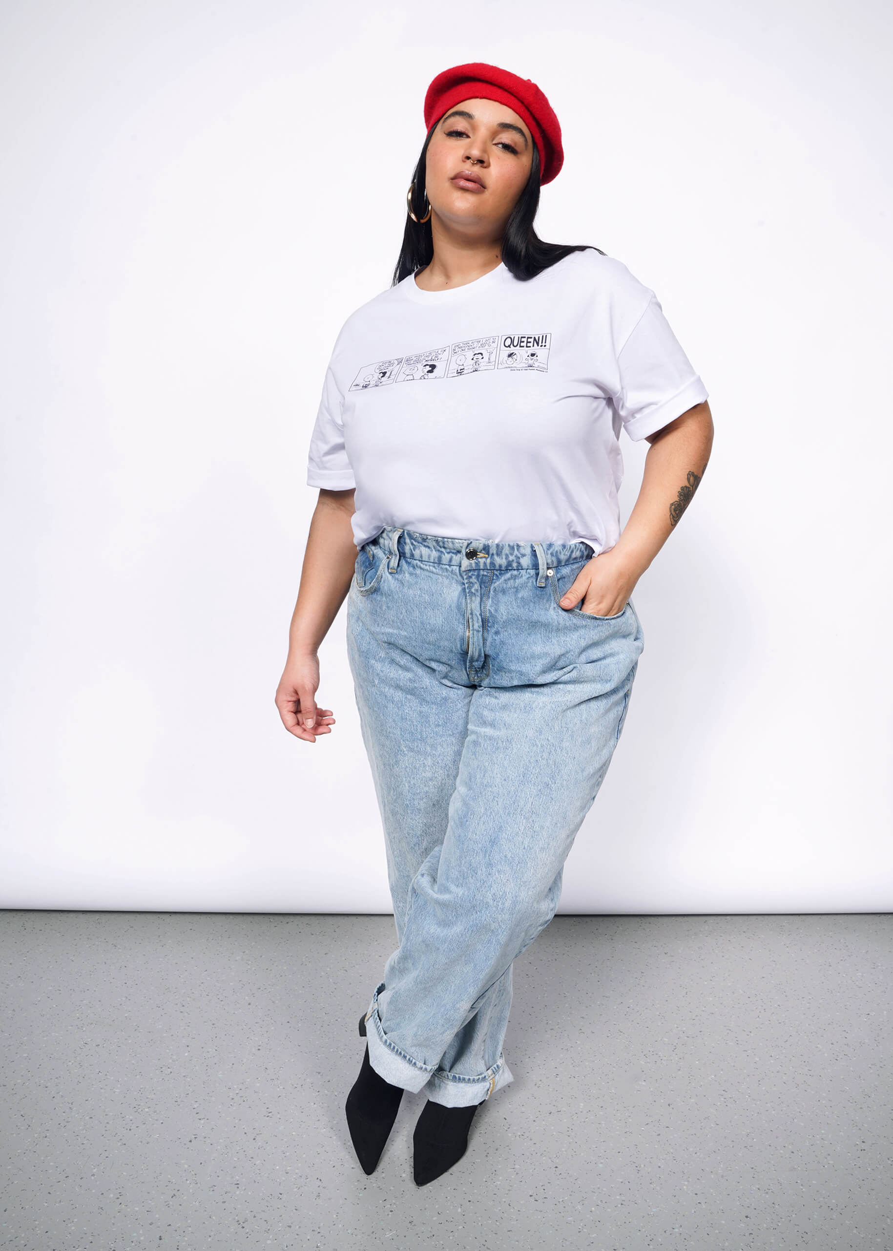 Person with long black hair looks at camera wearing tshirt with Peanuts comic strip printed on it, a red beret, jeans, and heeled boots.