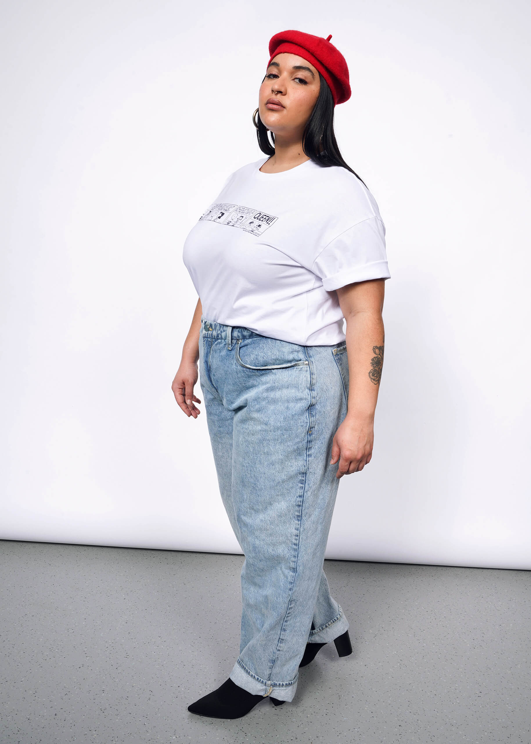 Side view of person with long black hair looking at camera wearing tshirt with Peanuts comic strip printed on it, a red beret, jeans, and heeled boots.