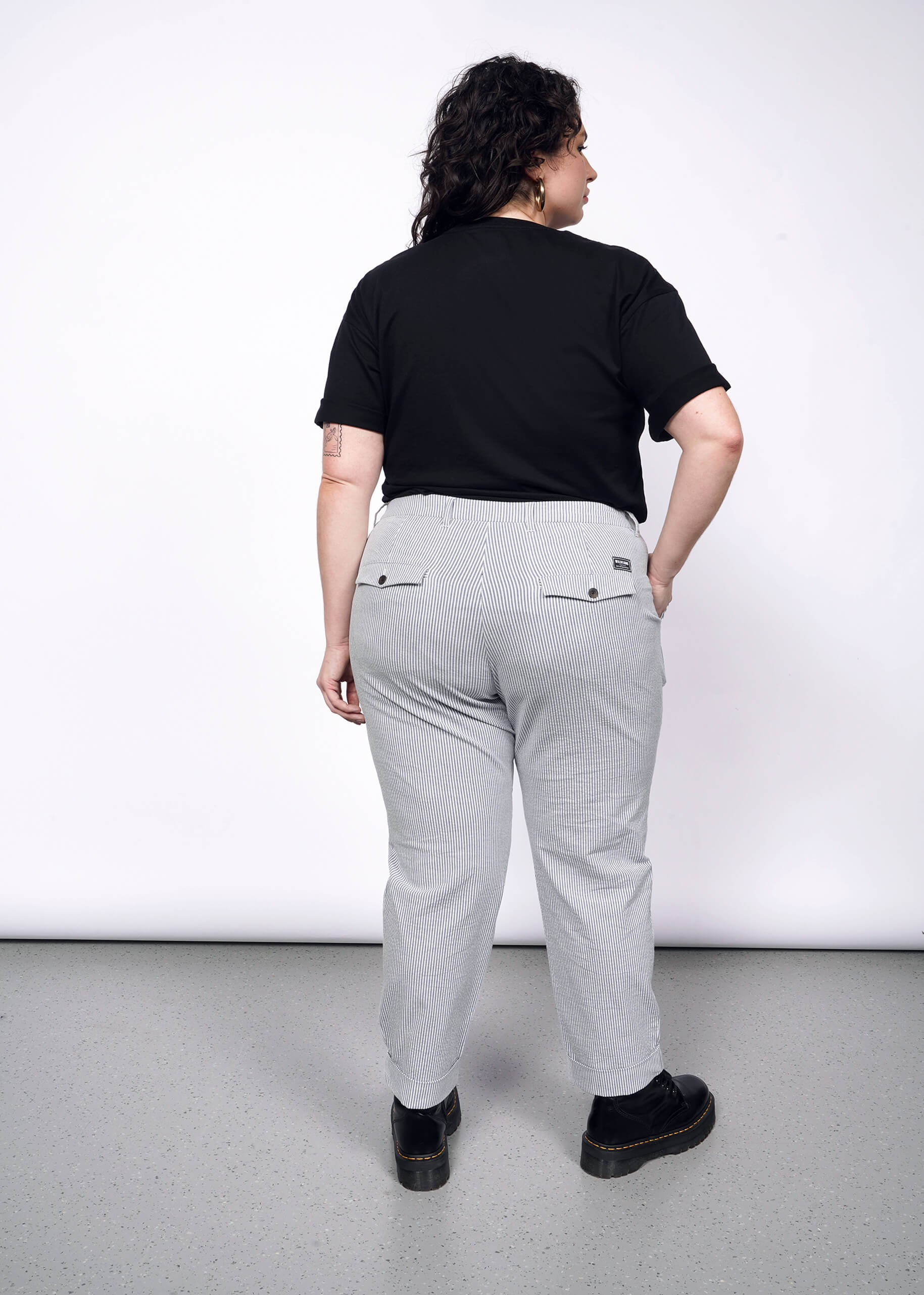 Back view of person with short curly black hair wearing hoop earrings, seersucker trousers, black tshirt, and black Doc Martens.