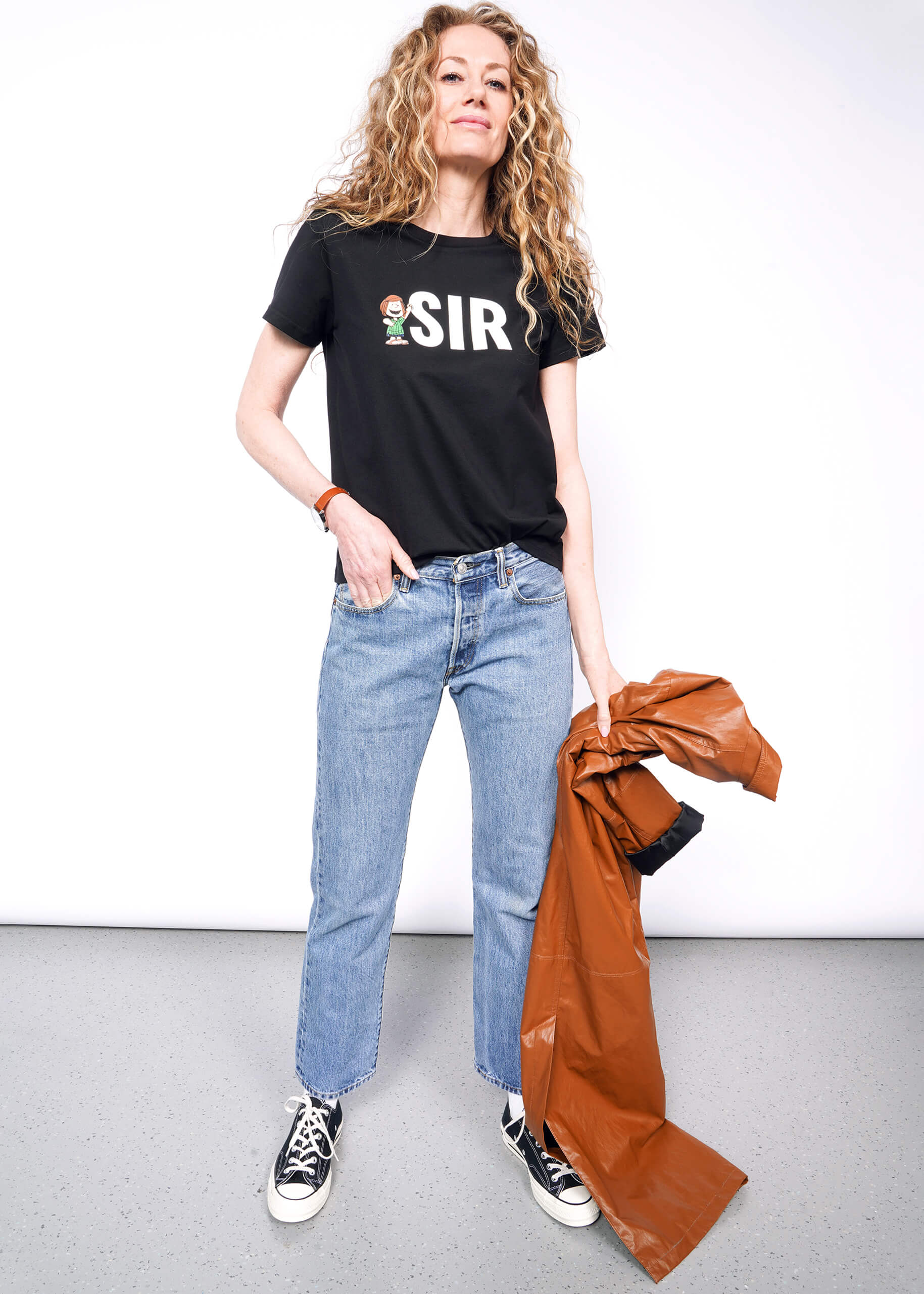 Person with wavy blonde hair looks at camera while holding brown jacket wearing black tshirt with "SIR" and Peppermint Patty printed on it, jeans, and black Converse.