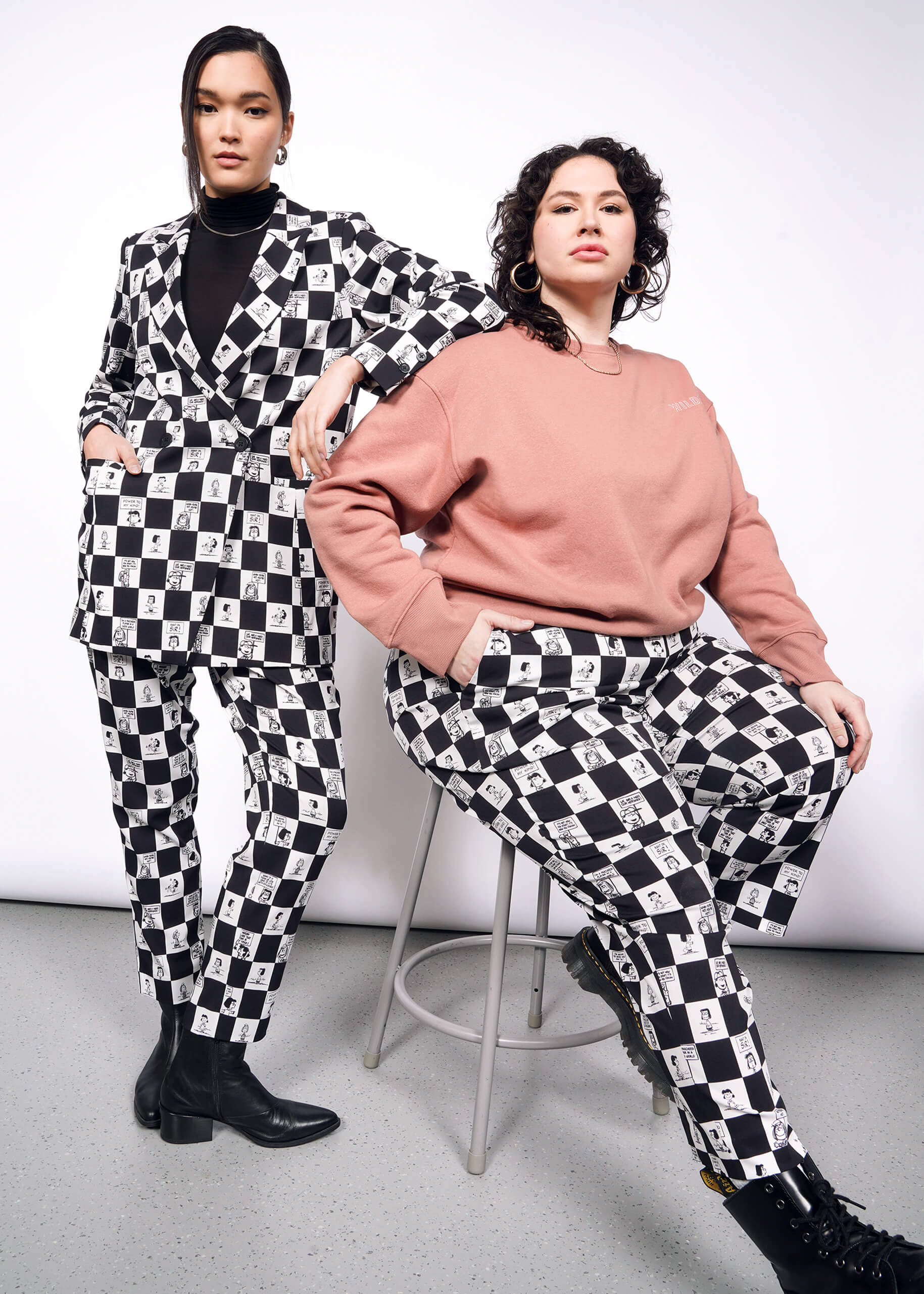 Person with black hair tied back wears the checkered black and white Slim Crop Pant and Double Breasted Blazer from the Peanuts x Wildfang collection while standing with one hand on shoulder of sitting model. Other model, with short curly black hair, is sitting on stool wearing pink sweatshirt and the checkered black and white Slim Crop Pant from the Peanuts x Wildfang collection.