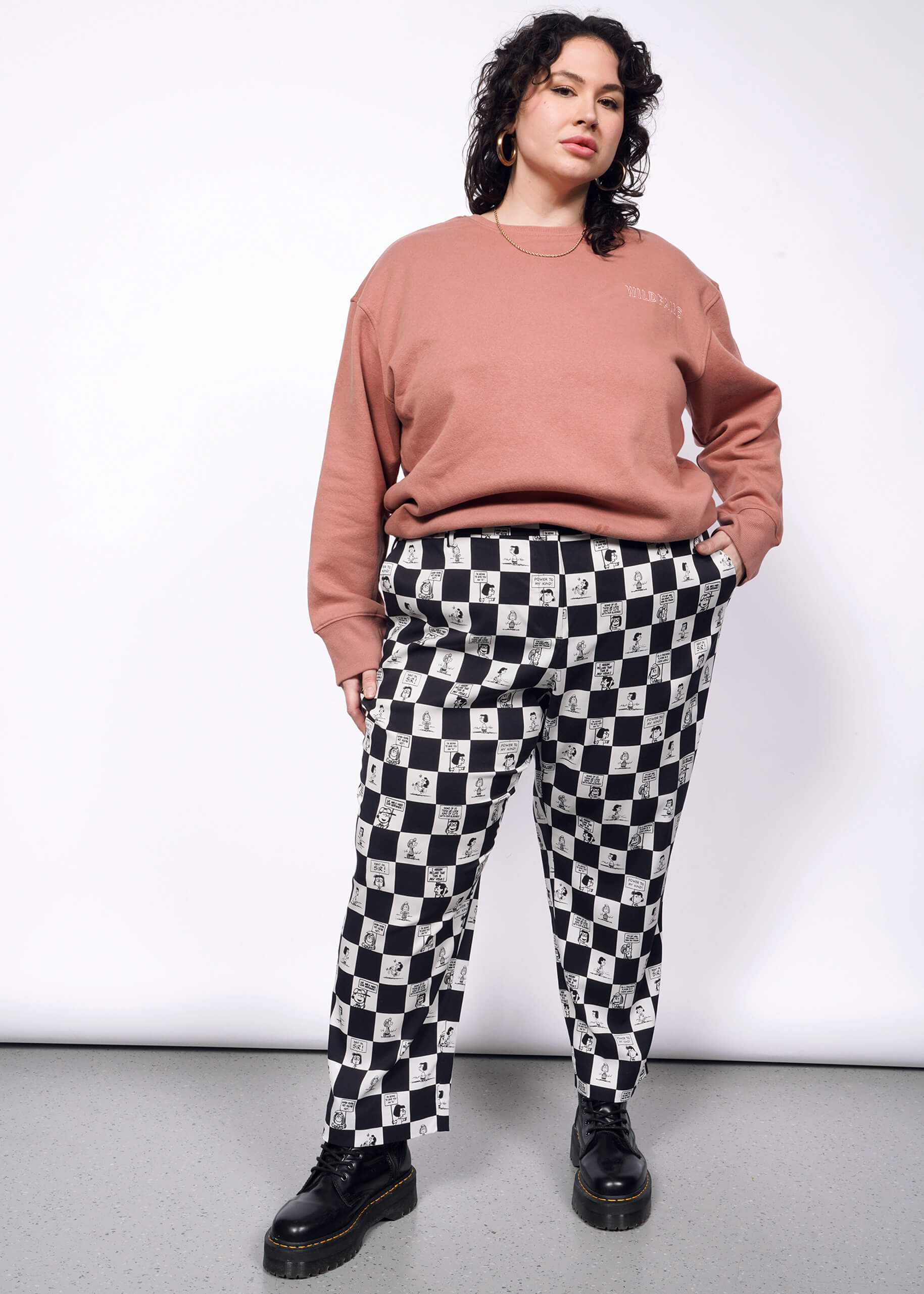 Person with short curly black hair looks at camera wearing pink sweatshirt, black boots, and the checkered black and white Slim Crop Pant from the Peanuts x Wildfang collection.