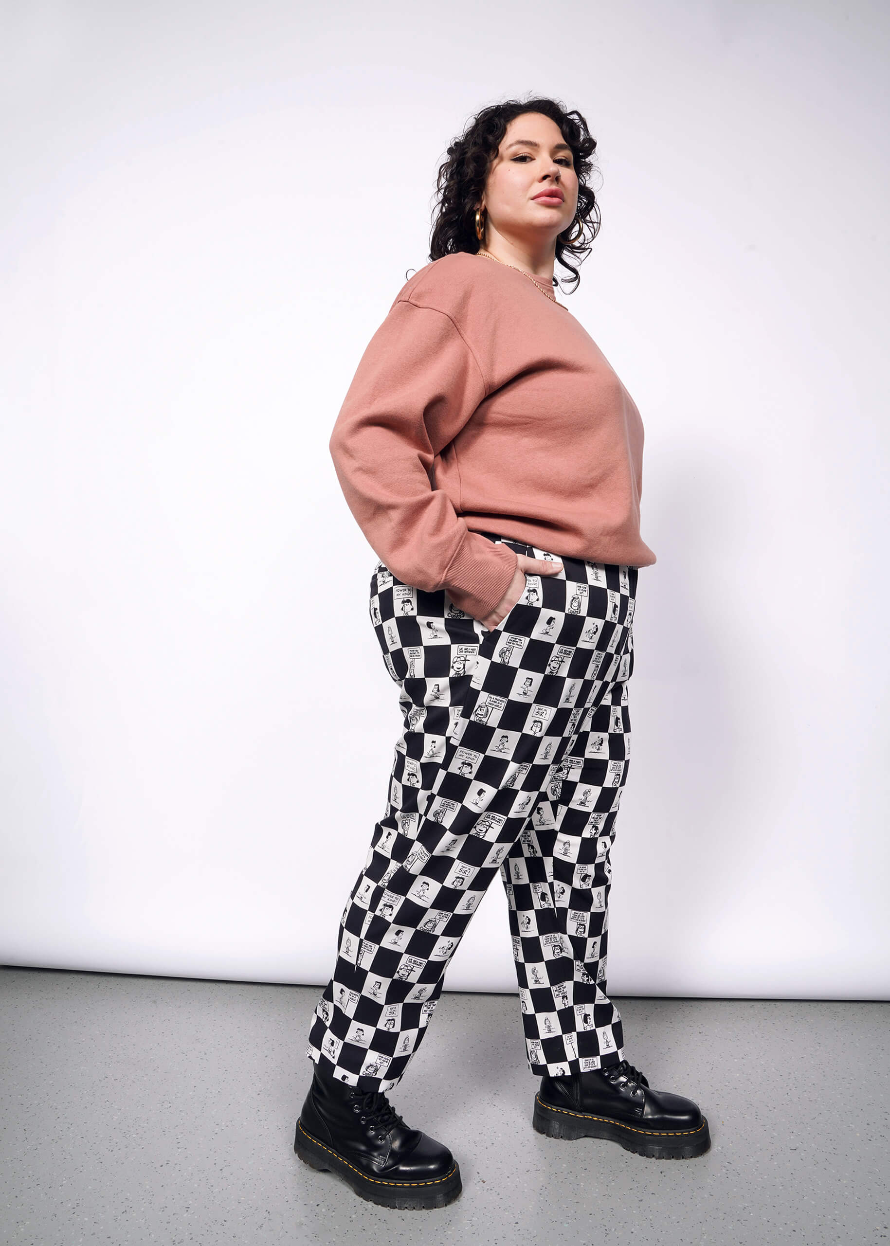 Side view of person with short curly black hair looking at camera with hand in pocket while wearing pink sweatshirt, black boots, and the checkered black and white Slim Crop Pant from the Peanuts x Wildfang collection.