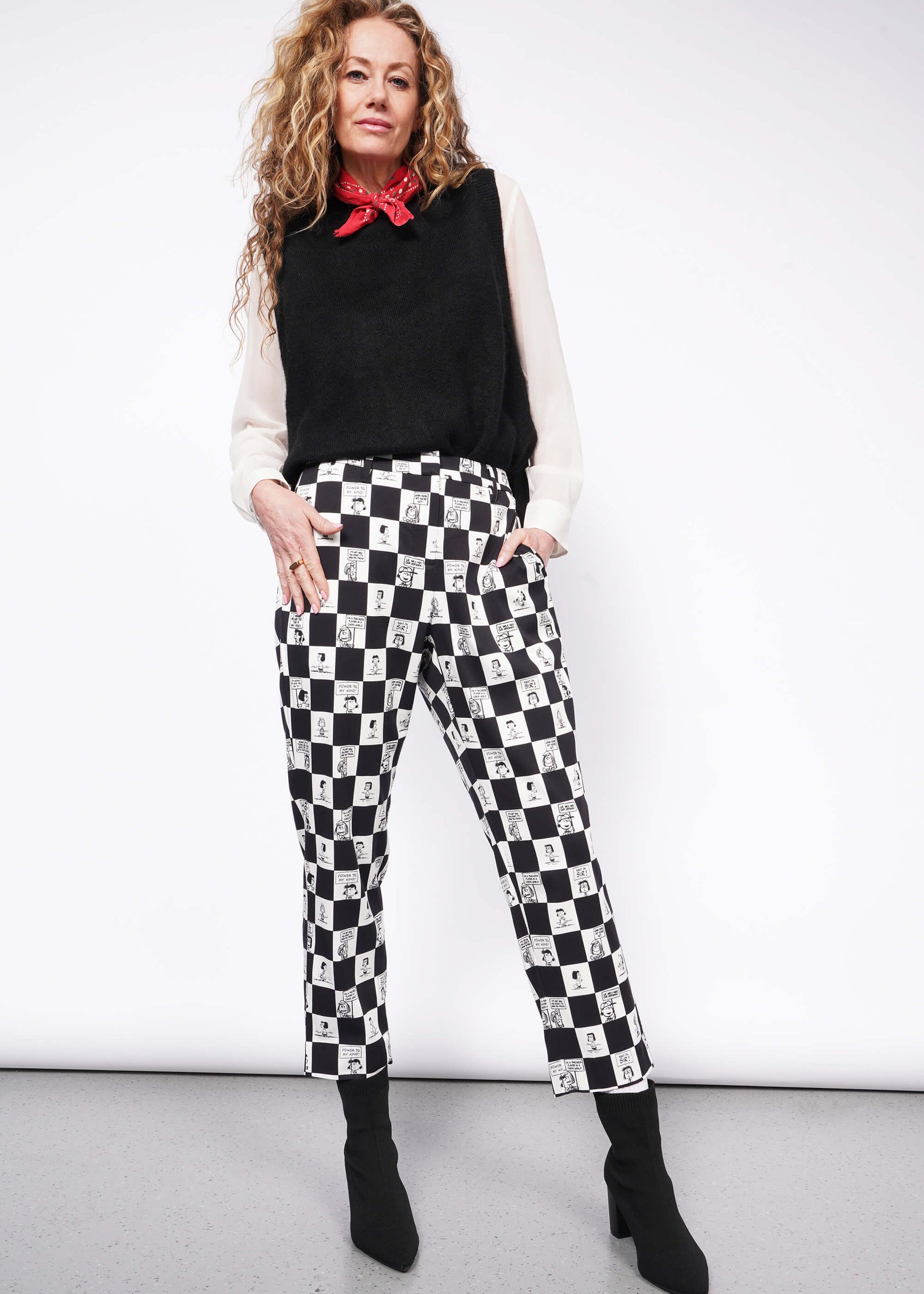 Person with wavy blonde hair looks at camera wearing white long sleeve button up under black sweater vest, a red bandana tied around their neck, black heeled boots, and the checkered black and white Slim Crop Pant from the Peanuts x Wildfang collection.