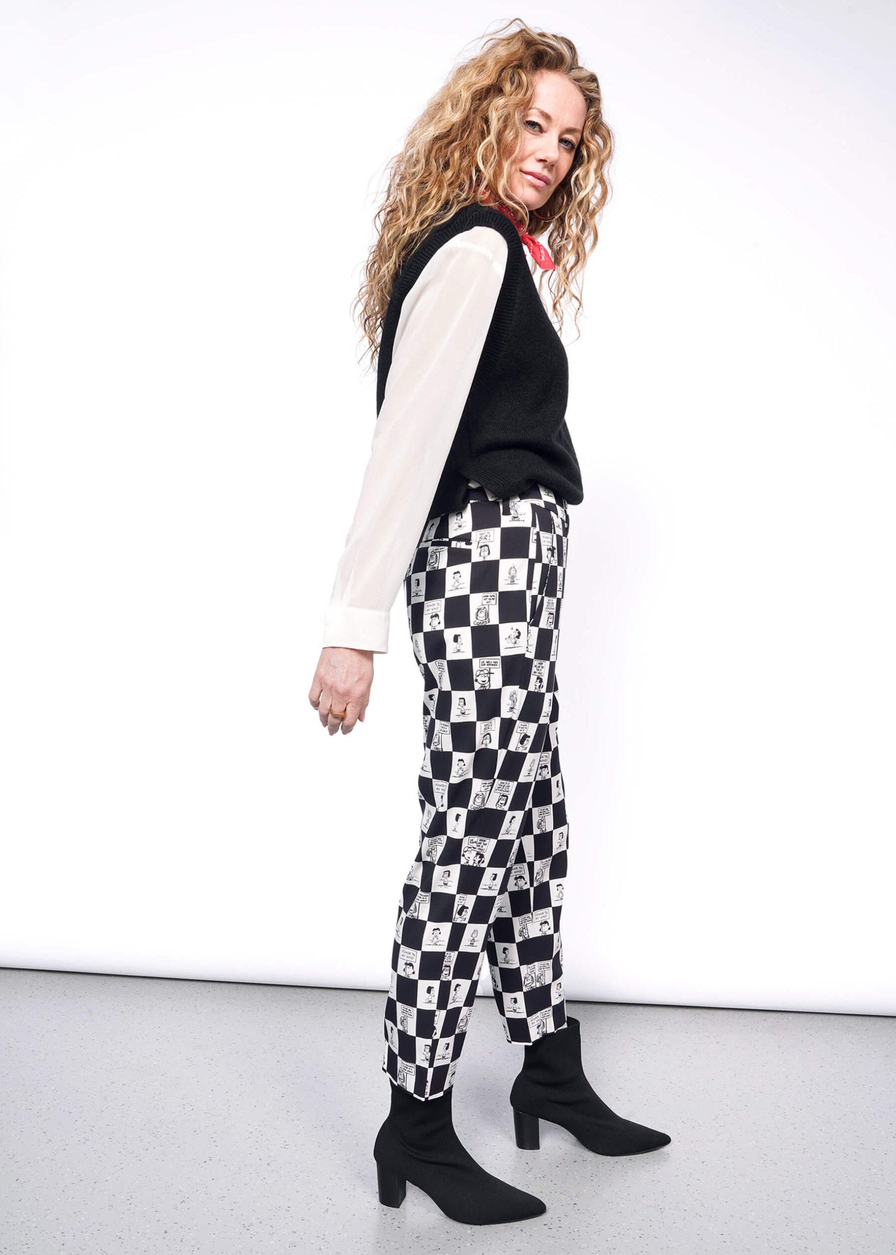 Side view of person with wavy blonde hair looking at camera wearing white long sleeve button up under black sweater vest, a red bandana tied around their neck, black heeled boots, and the checkered black and white Slim Crop Pant from the Peanuts x Wildfang collection.