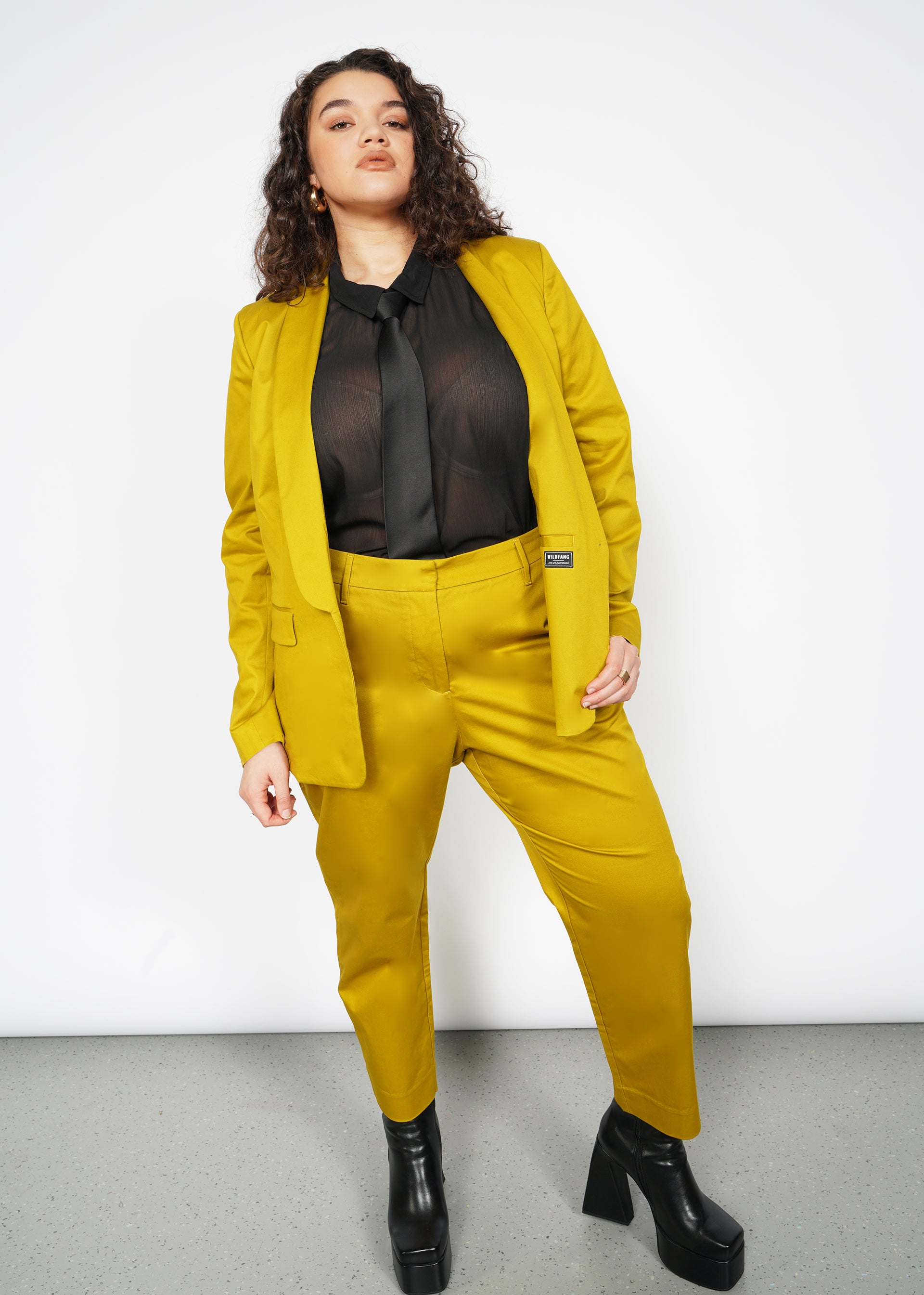 Confident person poses against a white background in a mustard yellow suit, featuring The Essential Slim Crop Pant with a tailored mid-rise fit. The ensemble is completed with a black sheer blouse, black tie, and chunky black boots.