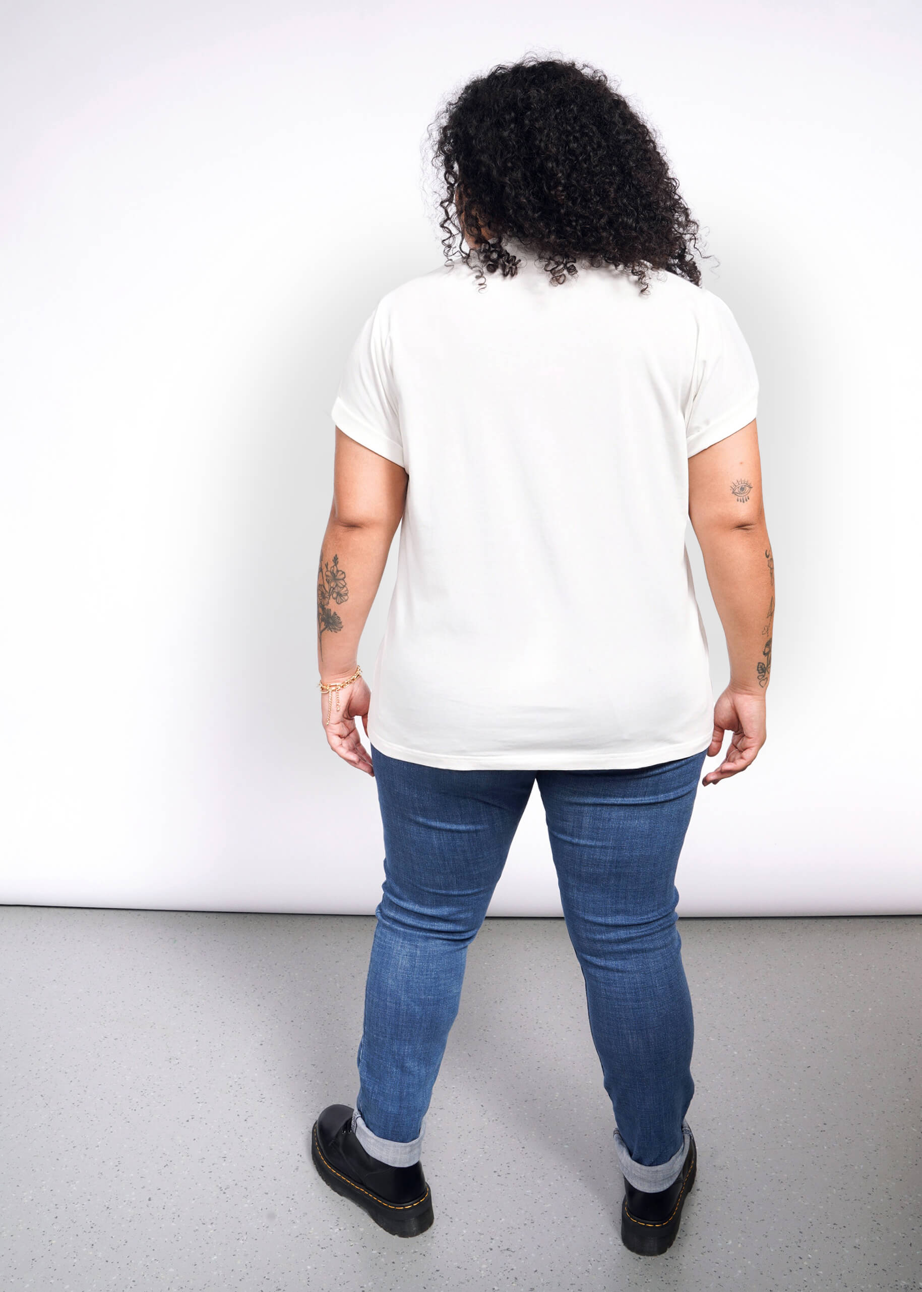 A curly-haired person, tattoos visible, stands back turned on a speckled gray floor. Theyre in a Debbie Harry X Wildfang Graphic Tee, blue jeans, and black shoes against a plain white backdrop, exuding punk fashion vibes.