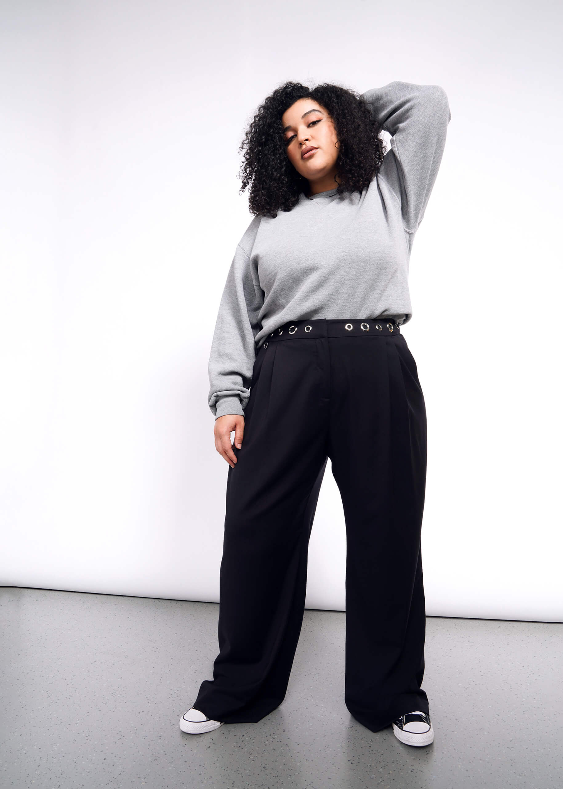 A person with curly hair poses confidently against a white backdrop, wearing a gray sweatshirt, Debbie Harry X Wildfang Wide Leg Trousers, and a studded belt. Black sneakers complete the look. One hand rests on their head as they exude a relaxed expression.