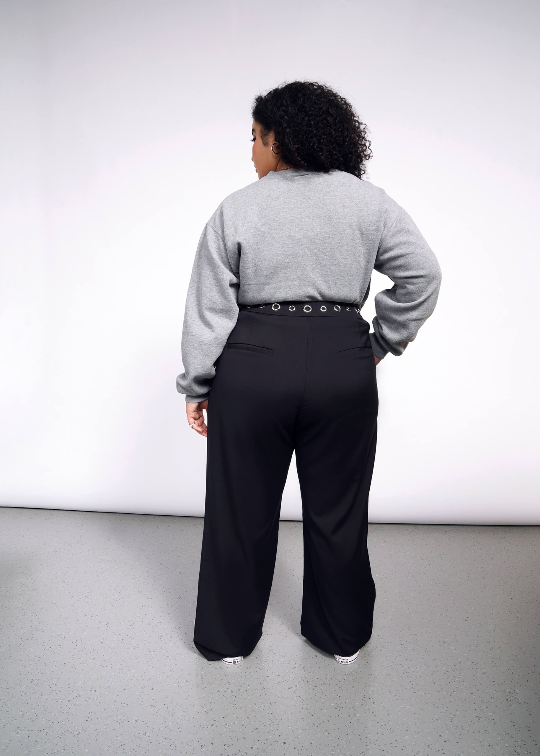 A person with curly hair faces away from the camera, wearing a gray sweater and black Debbie Harry X Wildfang Wide Leg Trouser. The minimalist backdrop features a gray floor, highlighting their chic ensemble.