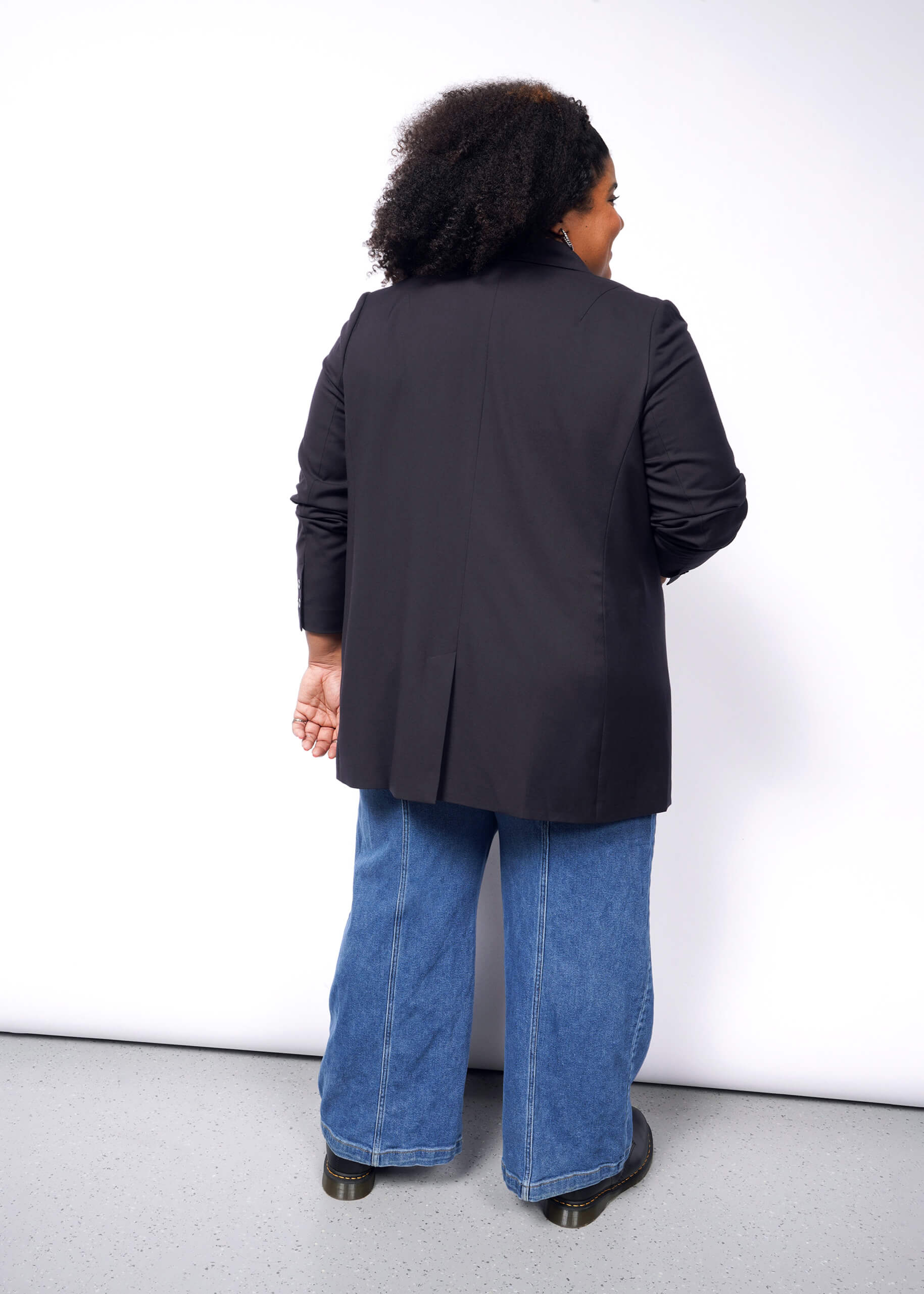 With their back to the camera, a person with curly hair wears The Empower Double Breasted Blazer and wide-leg blue jeans, exuding elegance against the plain white background.