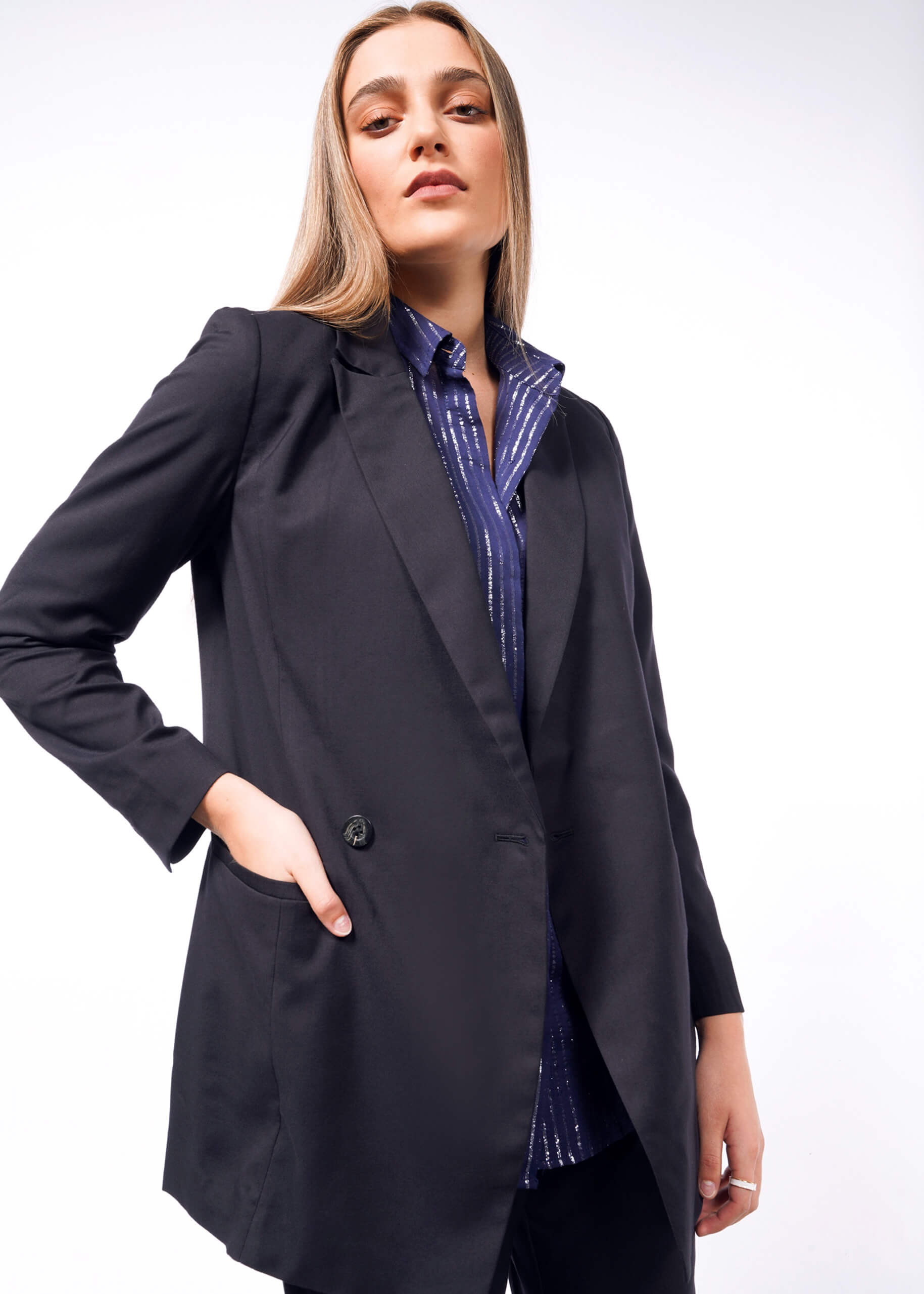 A person with long hair wears The Empower Double Breasted Blazer, tailored fit and dark, over a blue striped shirt, posing confidently with one hand in a pocket against a plain white background.