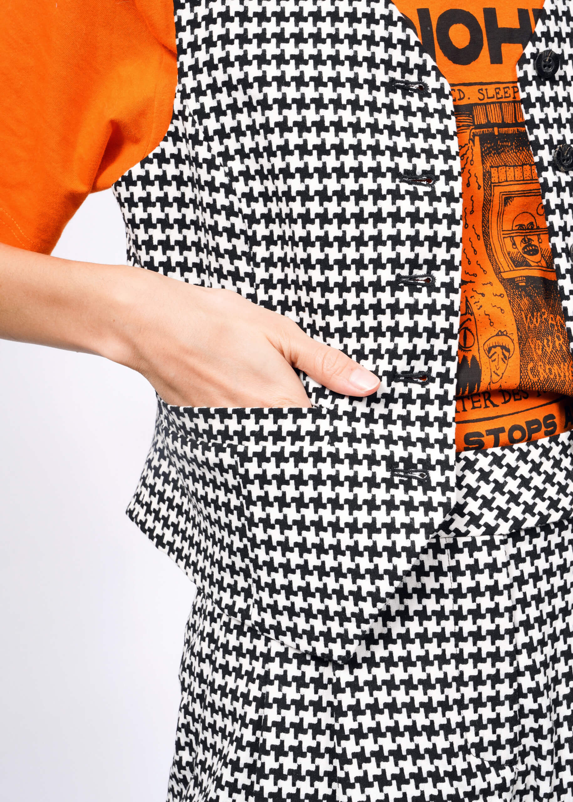 A close-up shows a person wearing The Empower Houndstooth Vest and pants with an orange graphic T-shirt beneath. Its ideal for versatile layering, as they casually have one hand in the vest pocket.