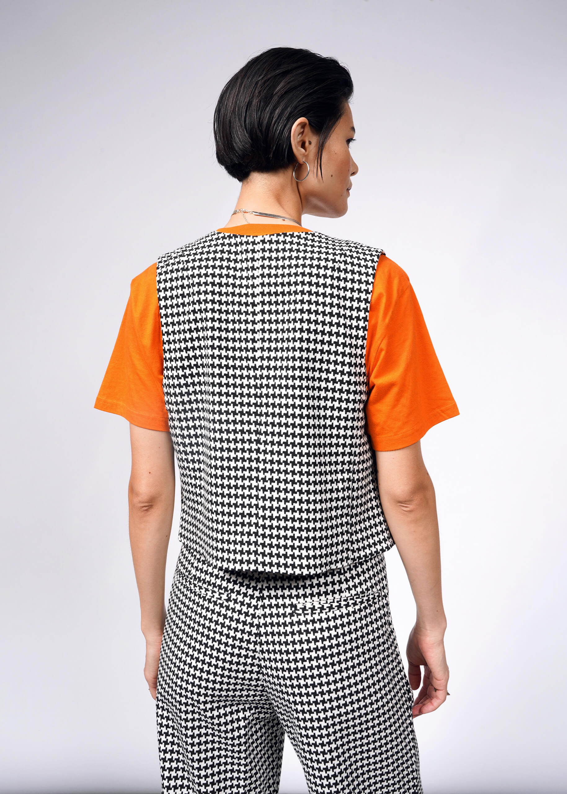 A person with short hair stands facing away, wearing The Empower Houndstooth Vest layered over an orange shirt, complemented by matching pants against a neutral gray background.