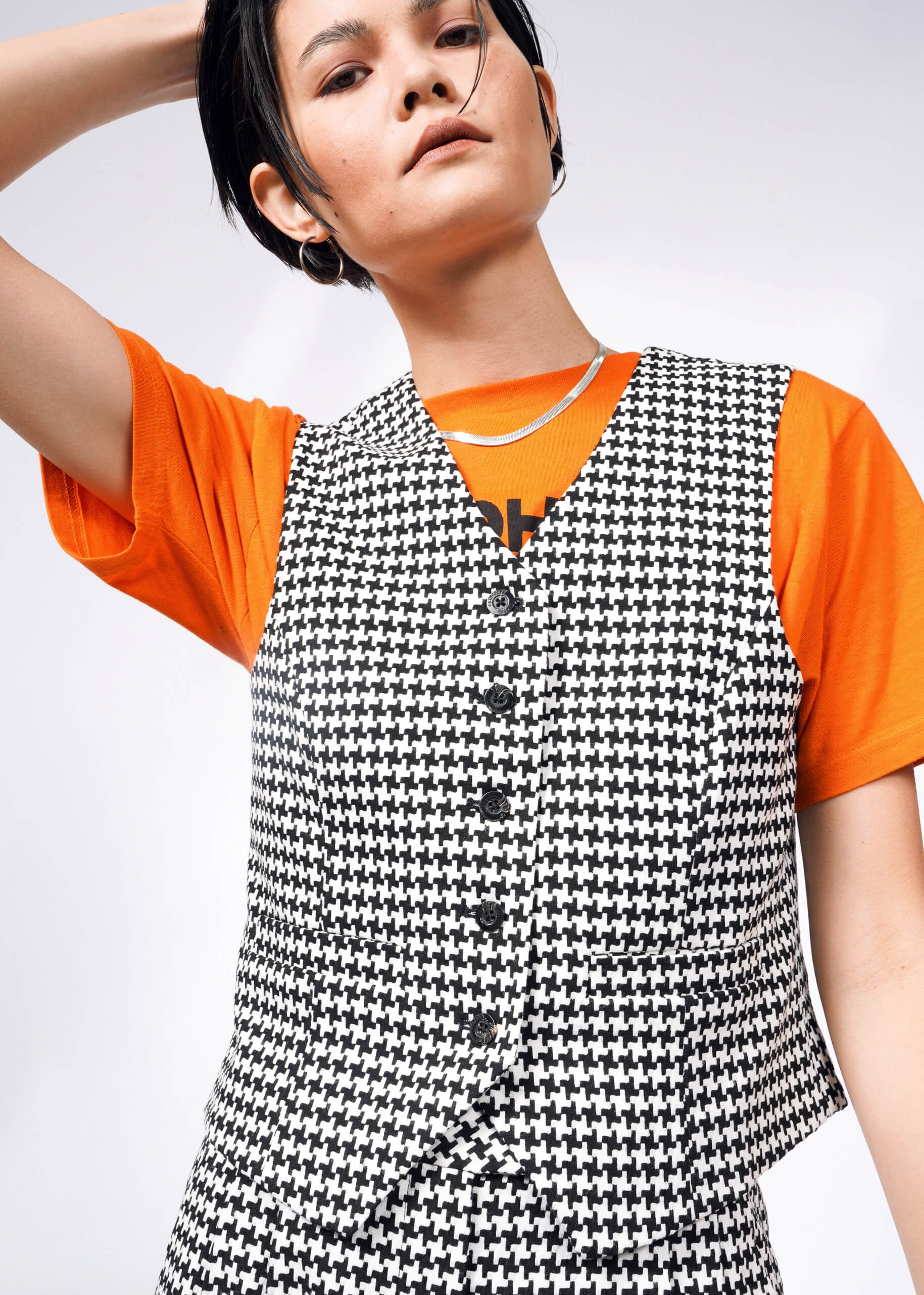 A person with short black hair wears an orange T-shirt beneath The Empower Houndstooth Vest. They pose with one arm raised against a plain white backdrop, showcasing this perfect wardrobe layering piece.