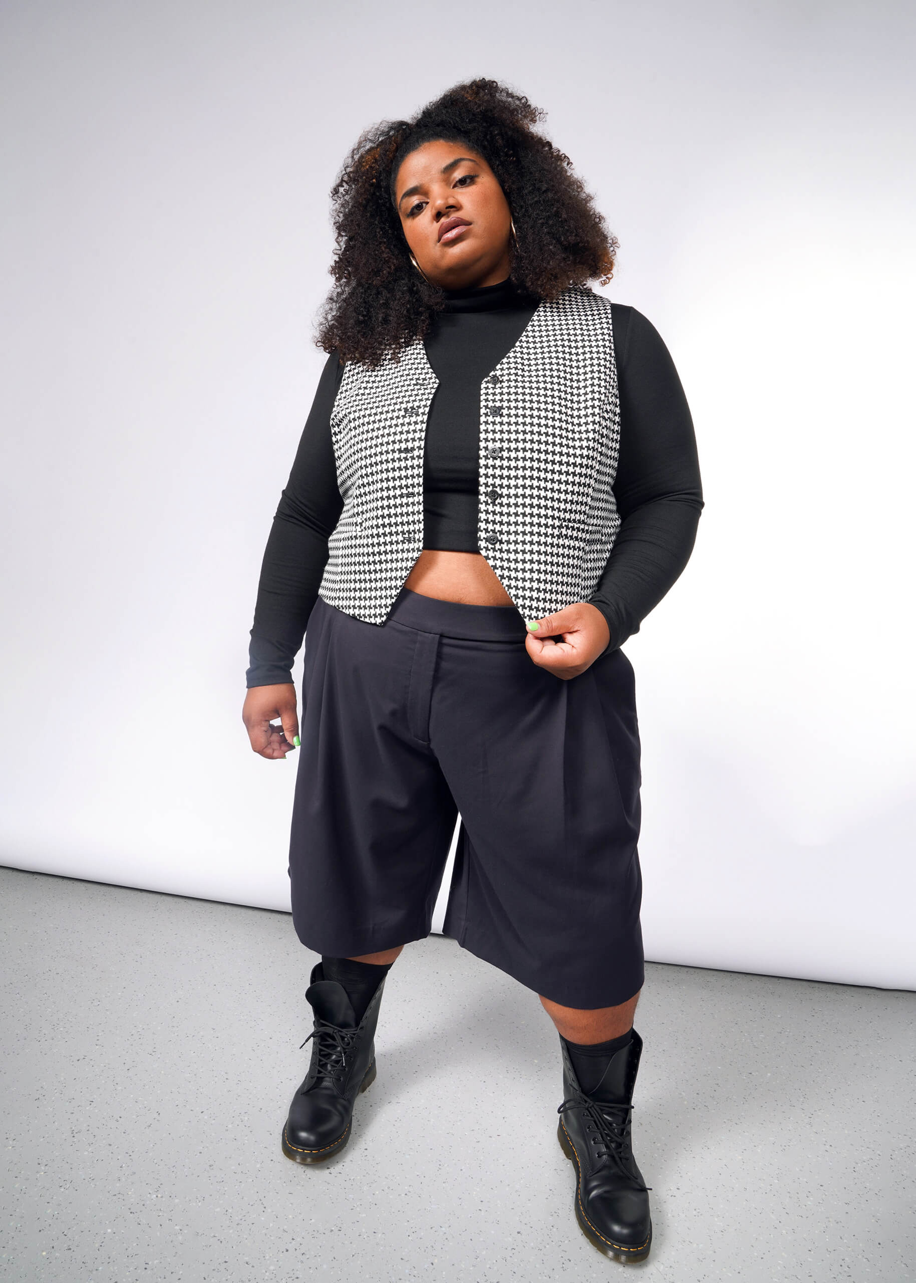 A curly-haired person exudes confidence against a neutral backdrop, dressed in a black turtleneck layered under The Empower Houndstooth Vest. Long black shorts and boots enhance their assertive pose and relaxed expression, completing the look.