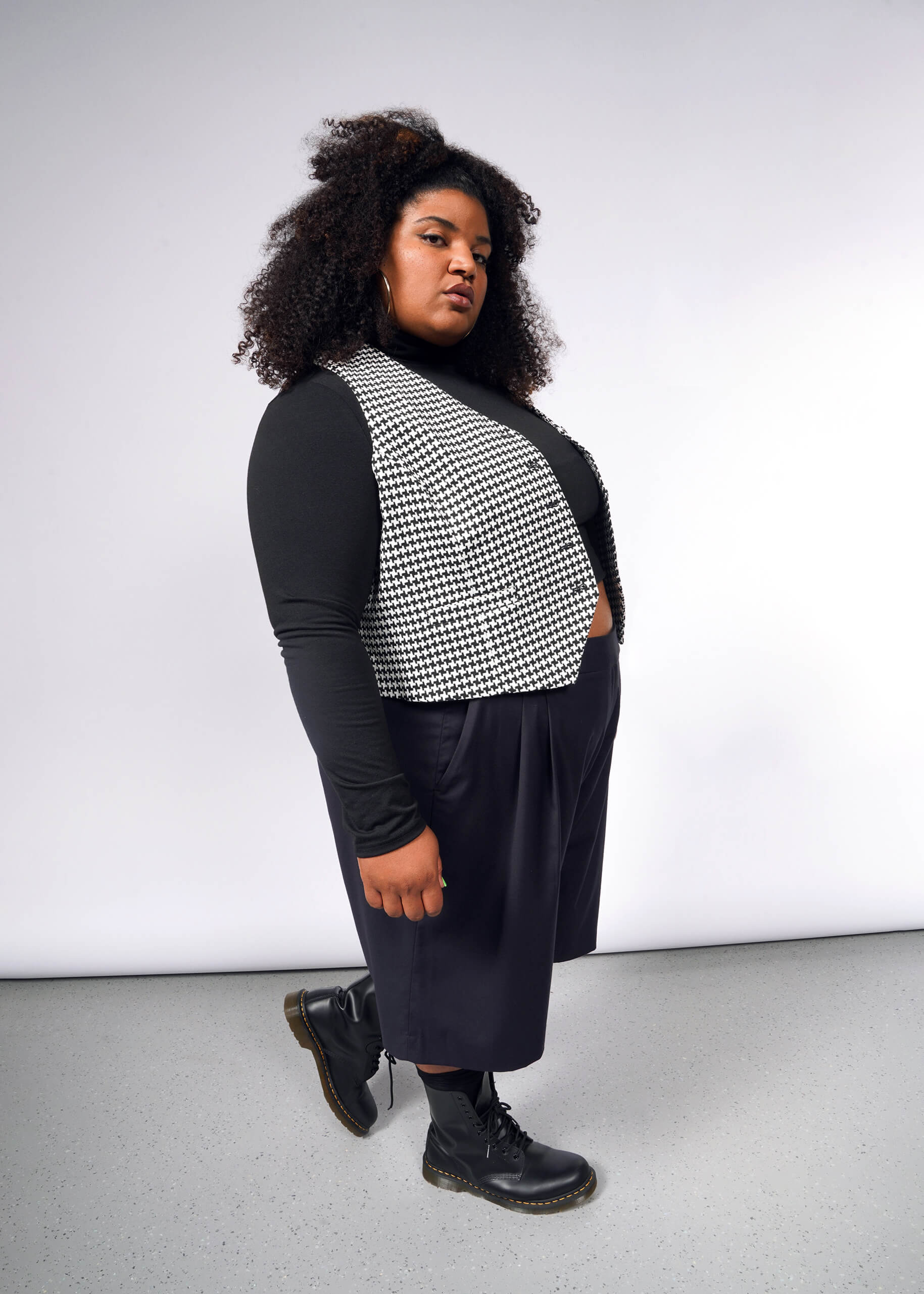 Against a plain, light background, a person with curly hair confidently wears a tailored black long-sleeve shirt, black skirt, and boots. They complete the ensemble with The Empower Houndstooth Vest as a stylish layering piece.