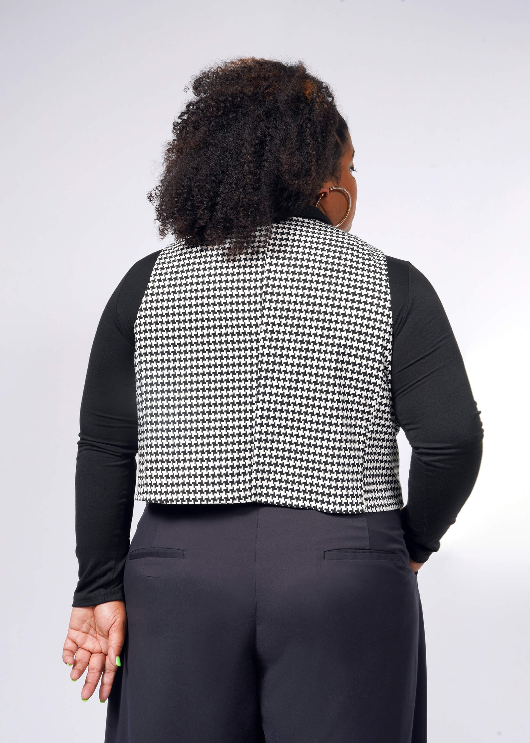 With curly hair facing away, someone highlights a tailored fit wearing The Empower Houndstooth Vest layered over a black long-sleeve top and matching pants. A plain, light background accentuates this stylish ensemble.