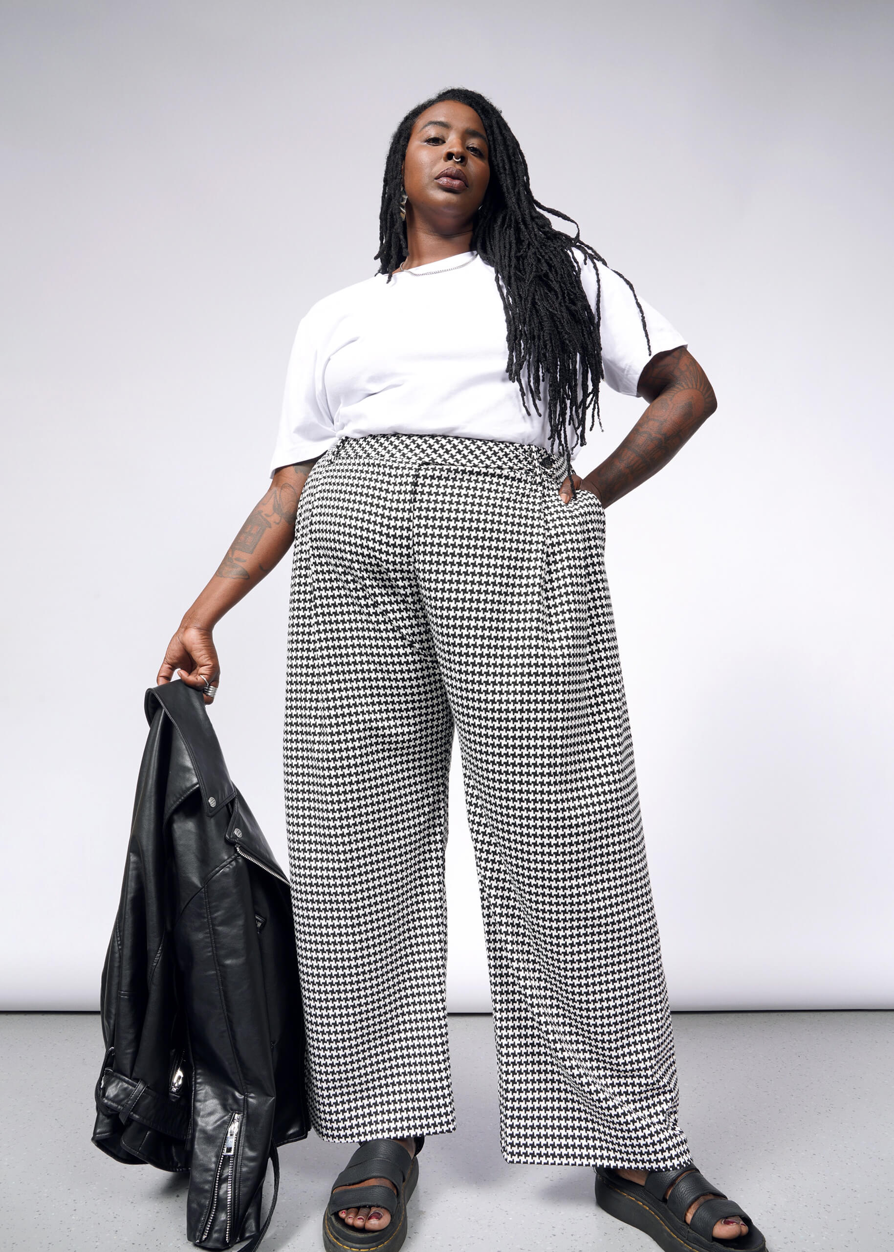 A person confidently poses against a plain background, wearing a white T-shirt and The Empower Houndstooth Wide Leg Trouser with double front pleats. They hold a black leather jacket in one hand and have long braided hair.