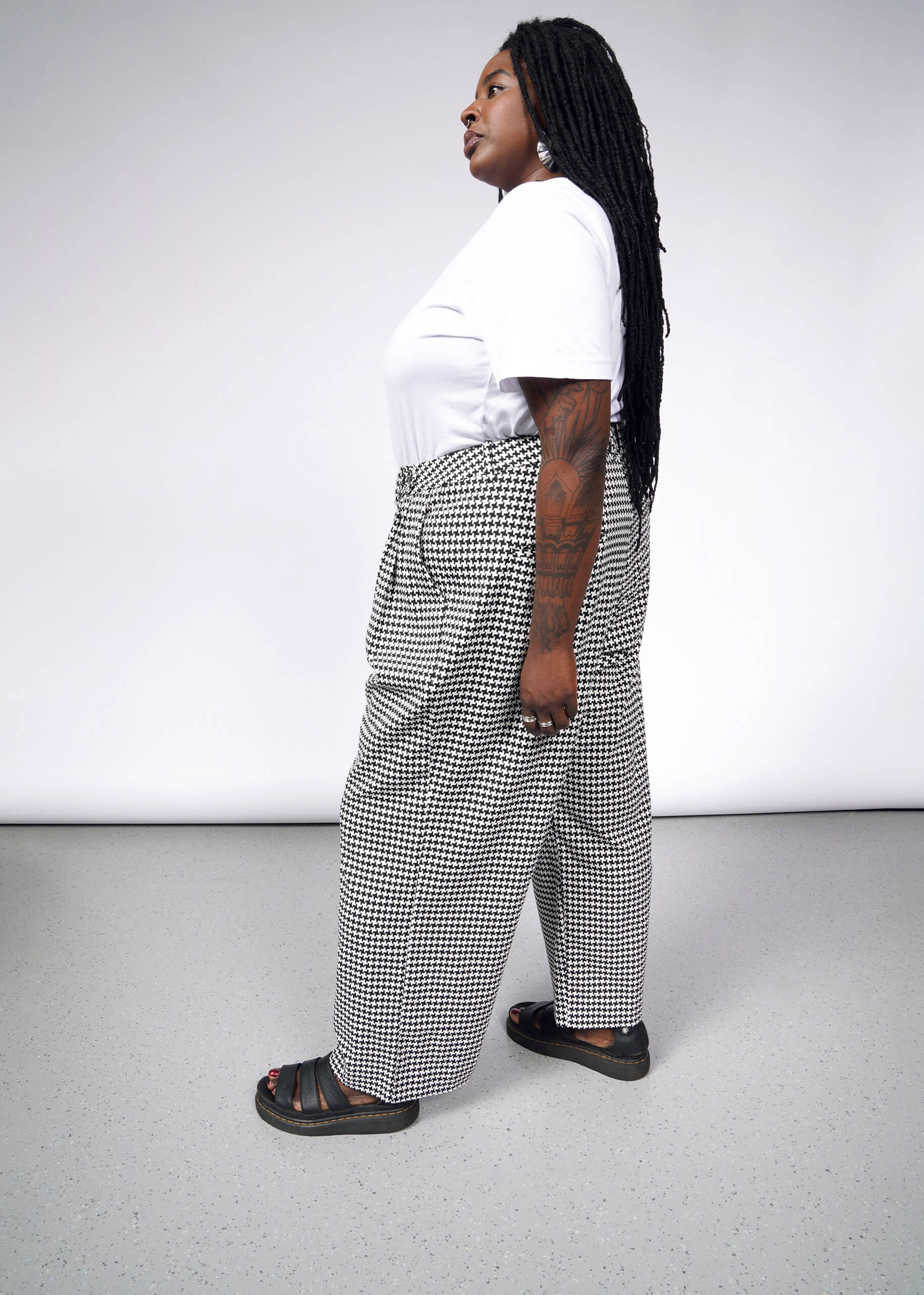 A person with long braided hair stands in profile against a plain background, wearing The Empower Houndstooth Wide Leg Trouser, a white t-shirt, black sandals, large hoop earrings, and displaying tattooed arms.