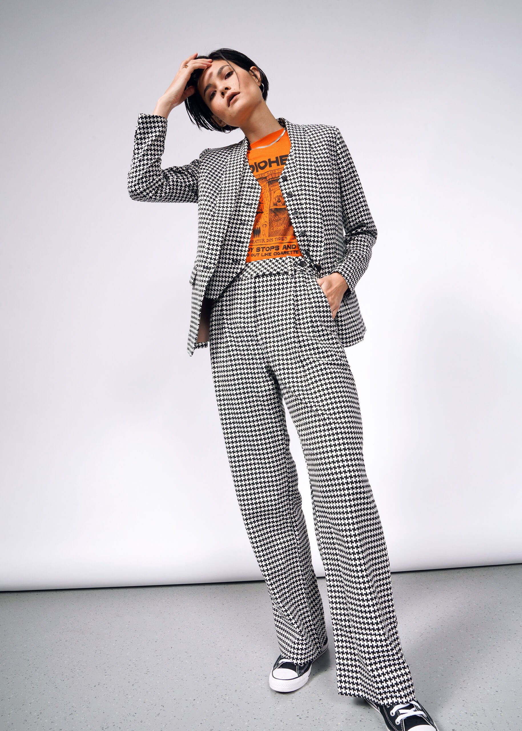 A person stands confidently in The Empower Houndstooth Wide Leg Trouser with double front pleats, paired with a bright orange graphic T-shirt and black Converse sneakers. One hand is in their pocket, and the other rests on their head against a plain background.