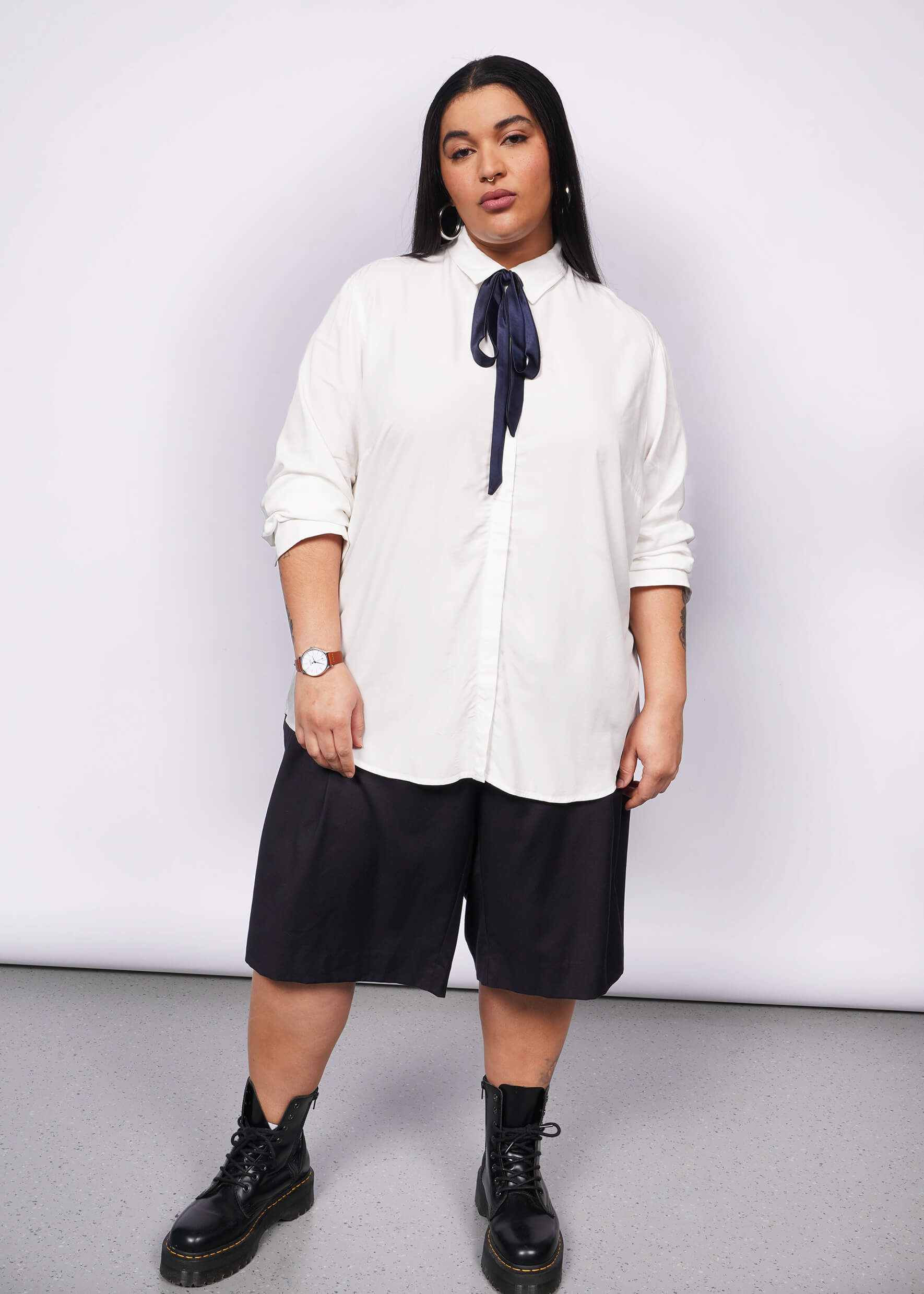 Person with long black hair looks into camera wearing long sleeve white button up with navy blue bow at collar, black wide leg shorts, and black boots.