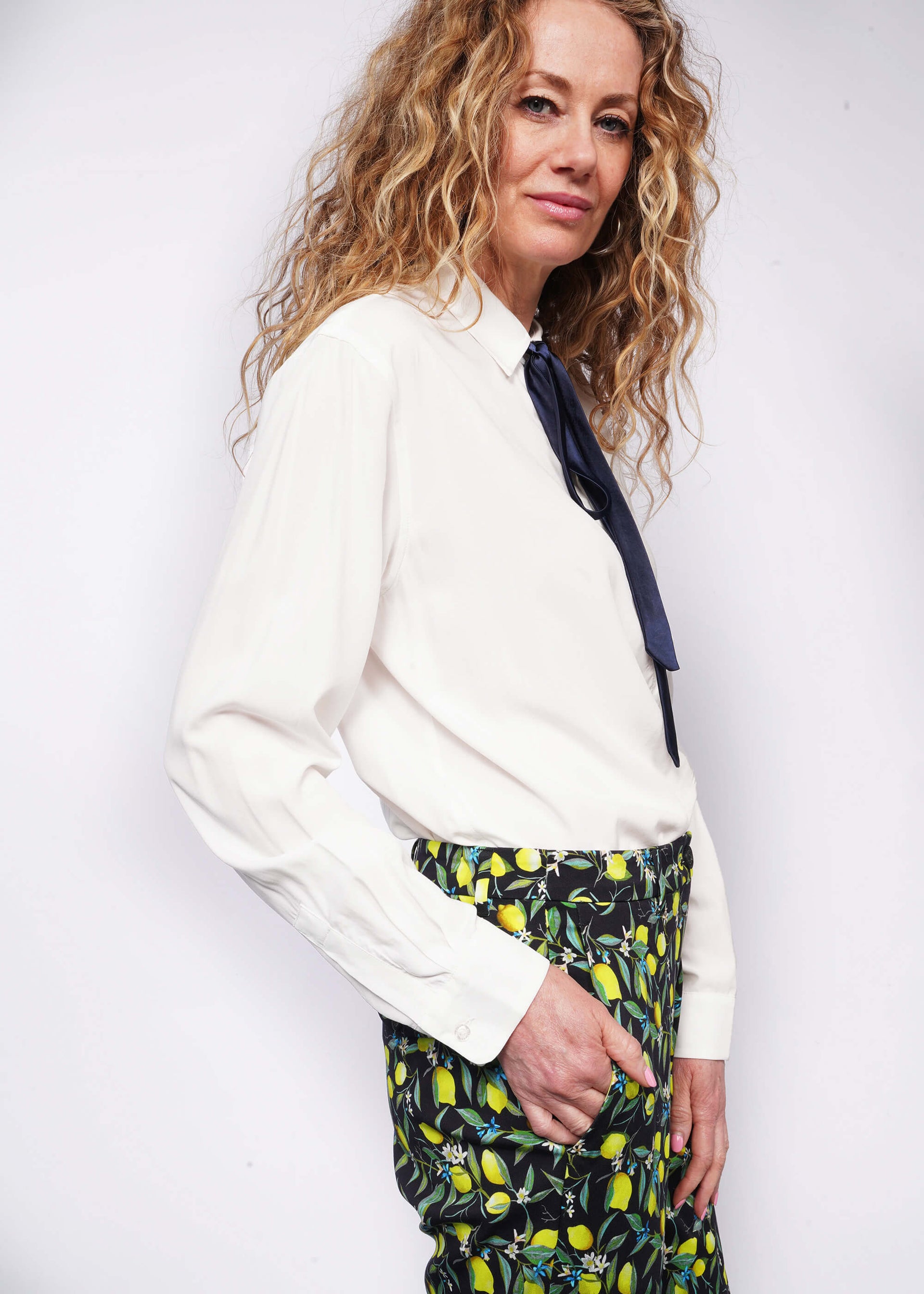 Side view of person with wavy blonde hair looking at camera with hand in pant pocket while wearing long sleeve white button up with bow at collar tucked into citrus printed trousers.