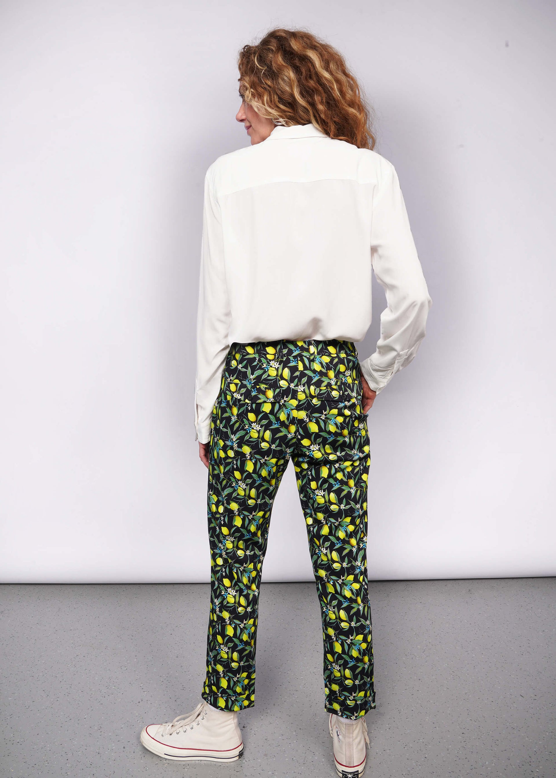 Back view of person with wavy blonde hair looking at camera with hand in pant pocket while wearing long sleeve white button up tucked into citrus printed trousers and Converse sneakers.