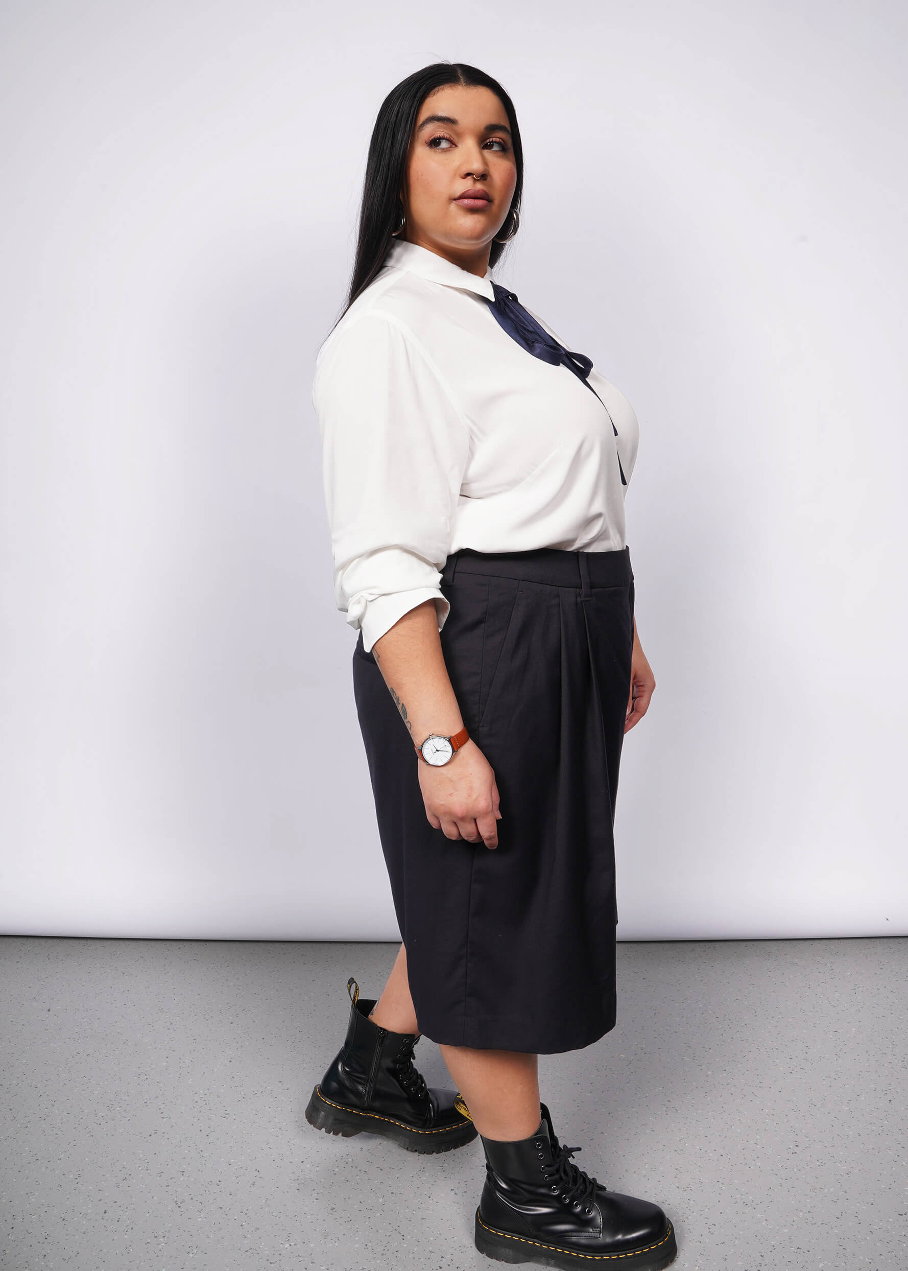 Side view of person with long black hair wears long sleeve white button up with navy blue bow at collar, black wide leg shorts, and black boots.