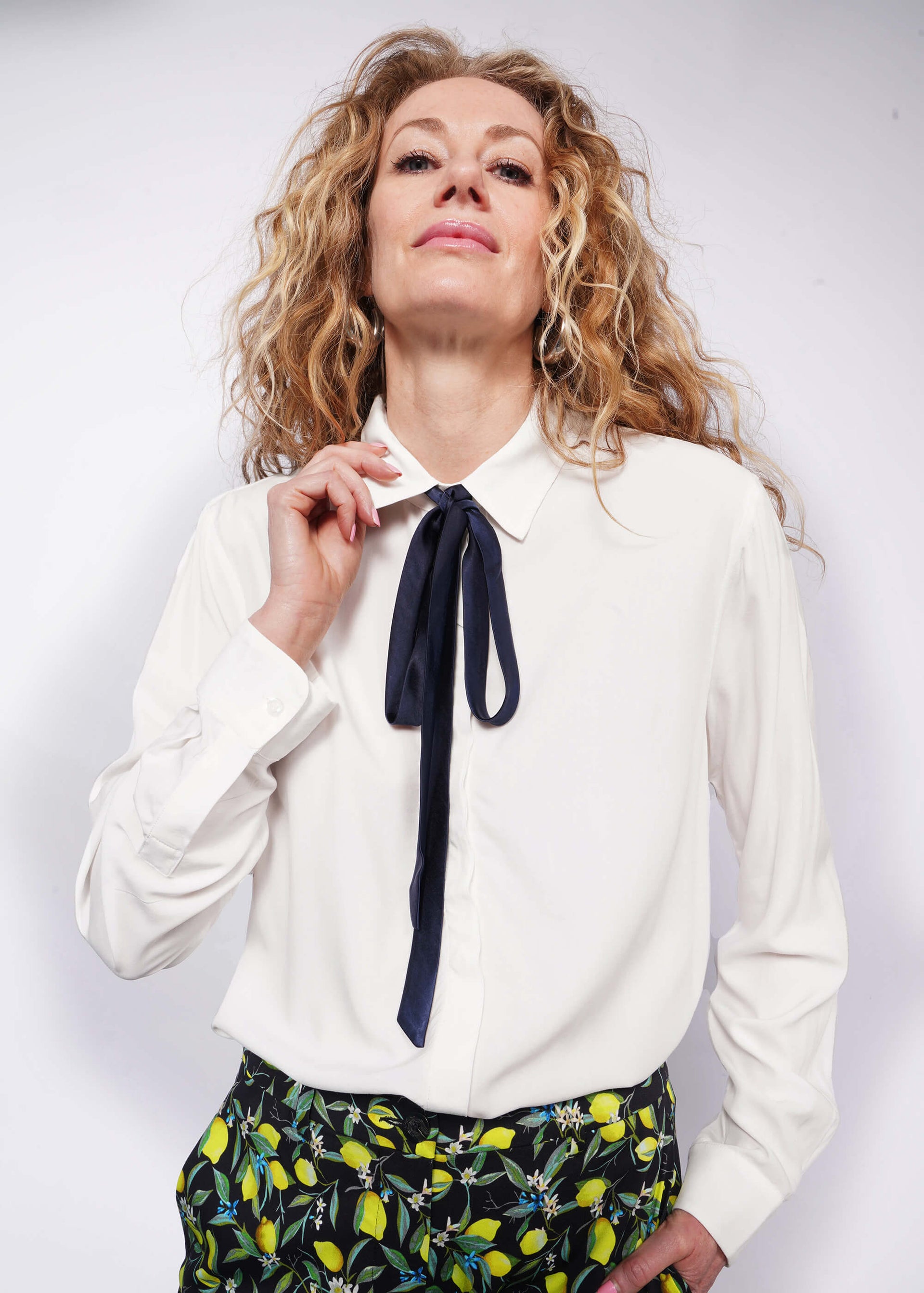 Person with wavy blonde hair looks at camera holding collar while wearing long sleeve white button up with bow at collar tucked into citrus printed trousers.