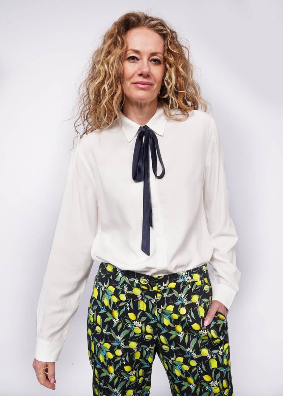 Person with wavy blonde hair looks at camera with hand in pant pocket while wearing long sleeve white button up with bow at collar tucked into citrus printed trousers.