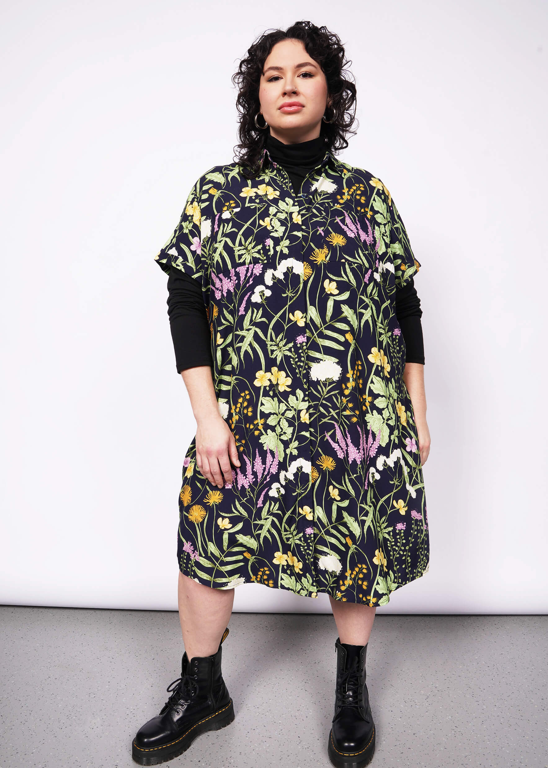 Person with short curly black hair looks at camera wearing black long sleeve turtleneck layered under short sleeved botanical printed shirt dress and Doc Martens boots.