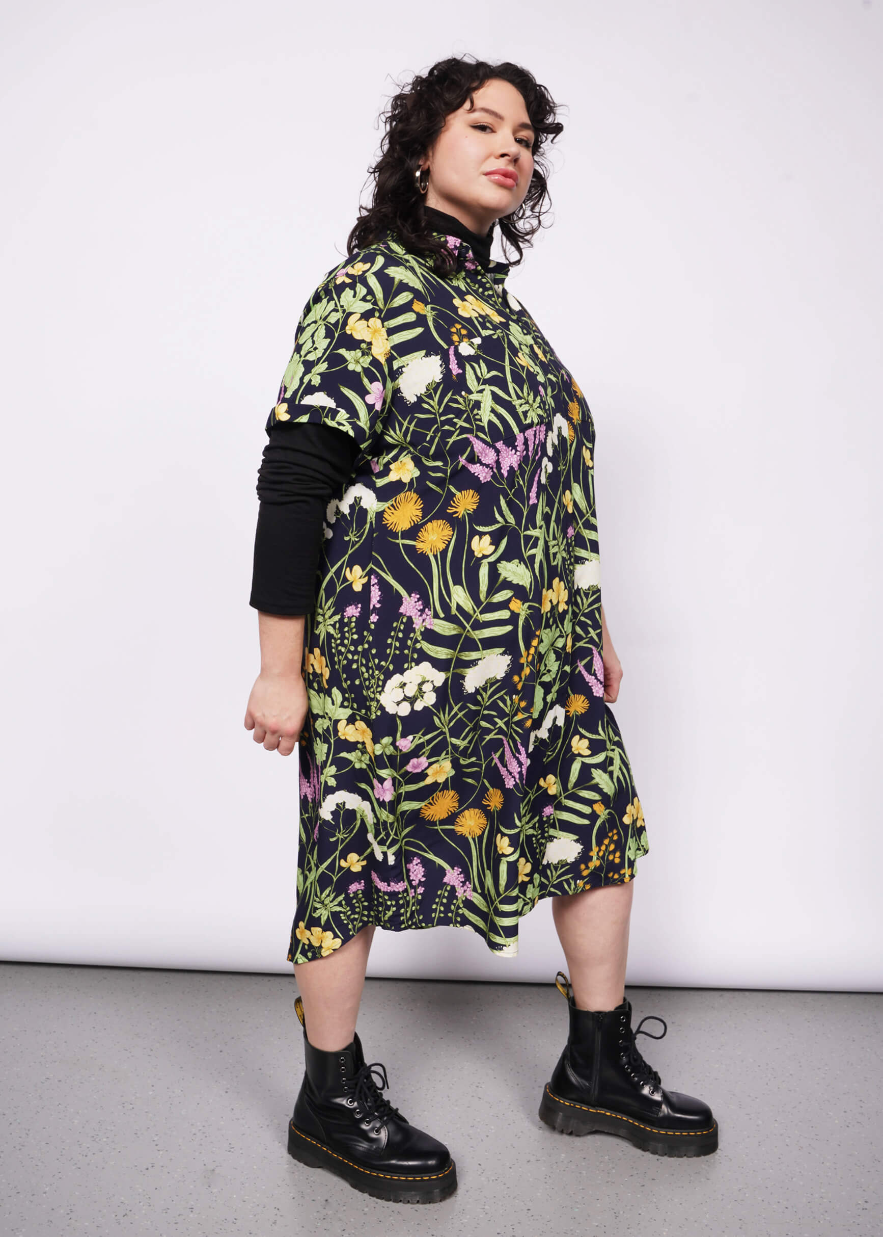 Side view of person with short curly black hair looking at camera wearing black long sleeve turtleneck layered under short sleeved botanical printed shirt dress and Doc Martens boots.