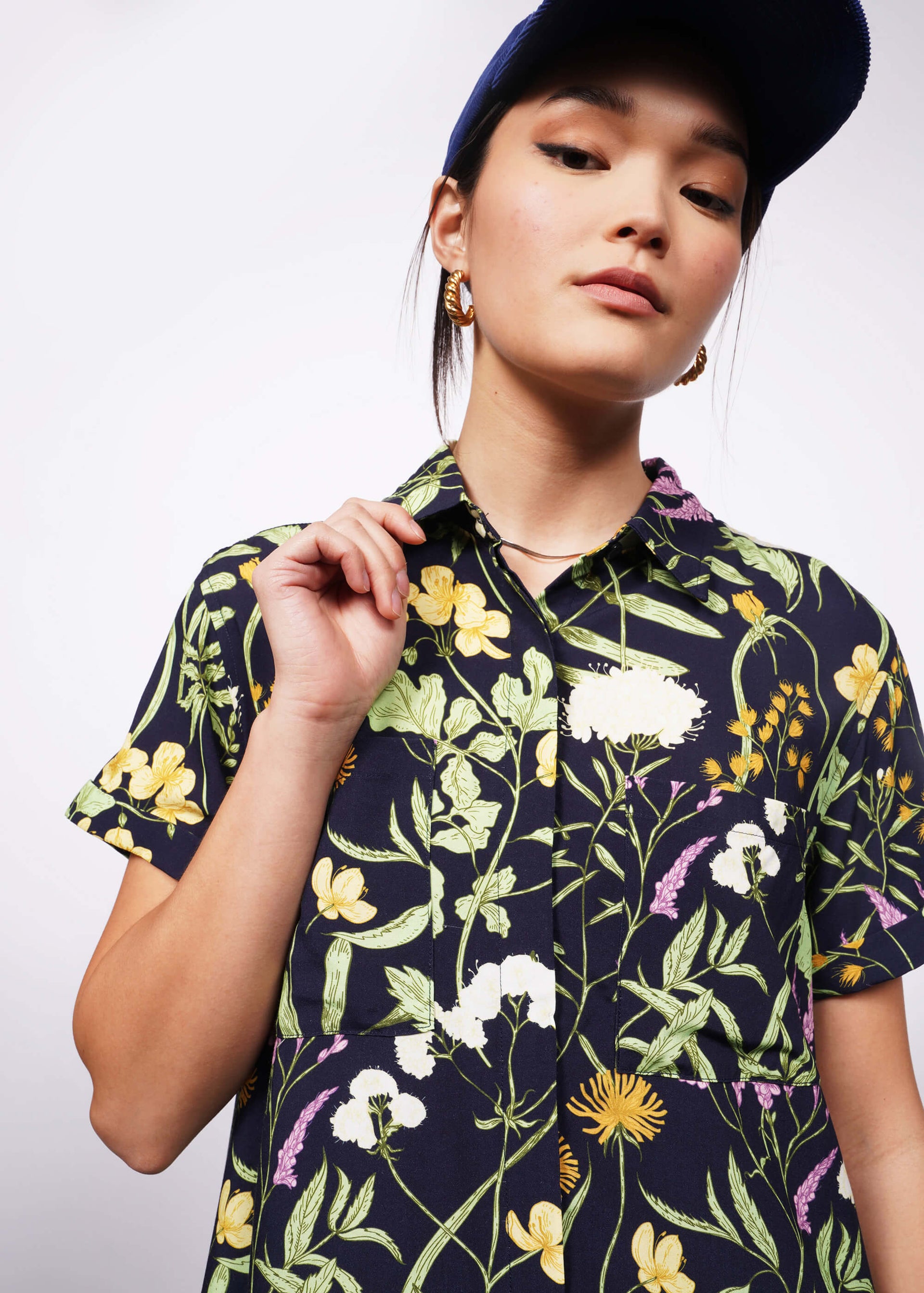 Close up view of person holding collar wearing botanical printed short sleeve button up shirt dress and baseball cap.