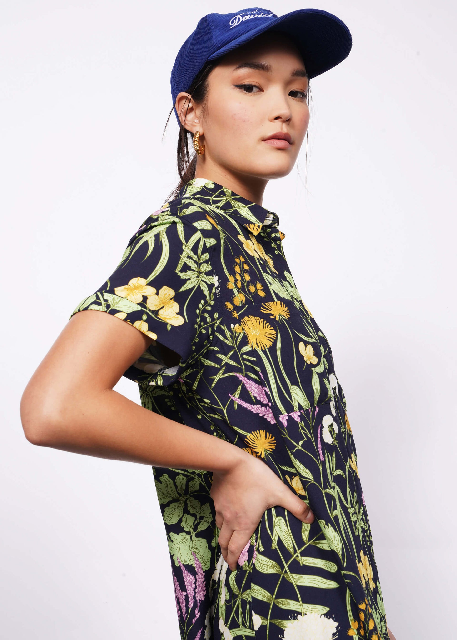 Close up of side view of person with hand on hip wearing baseball hat and botanical printed shirt dress.