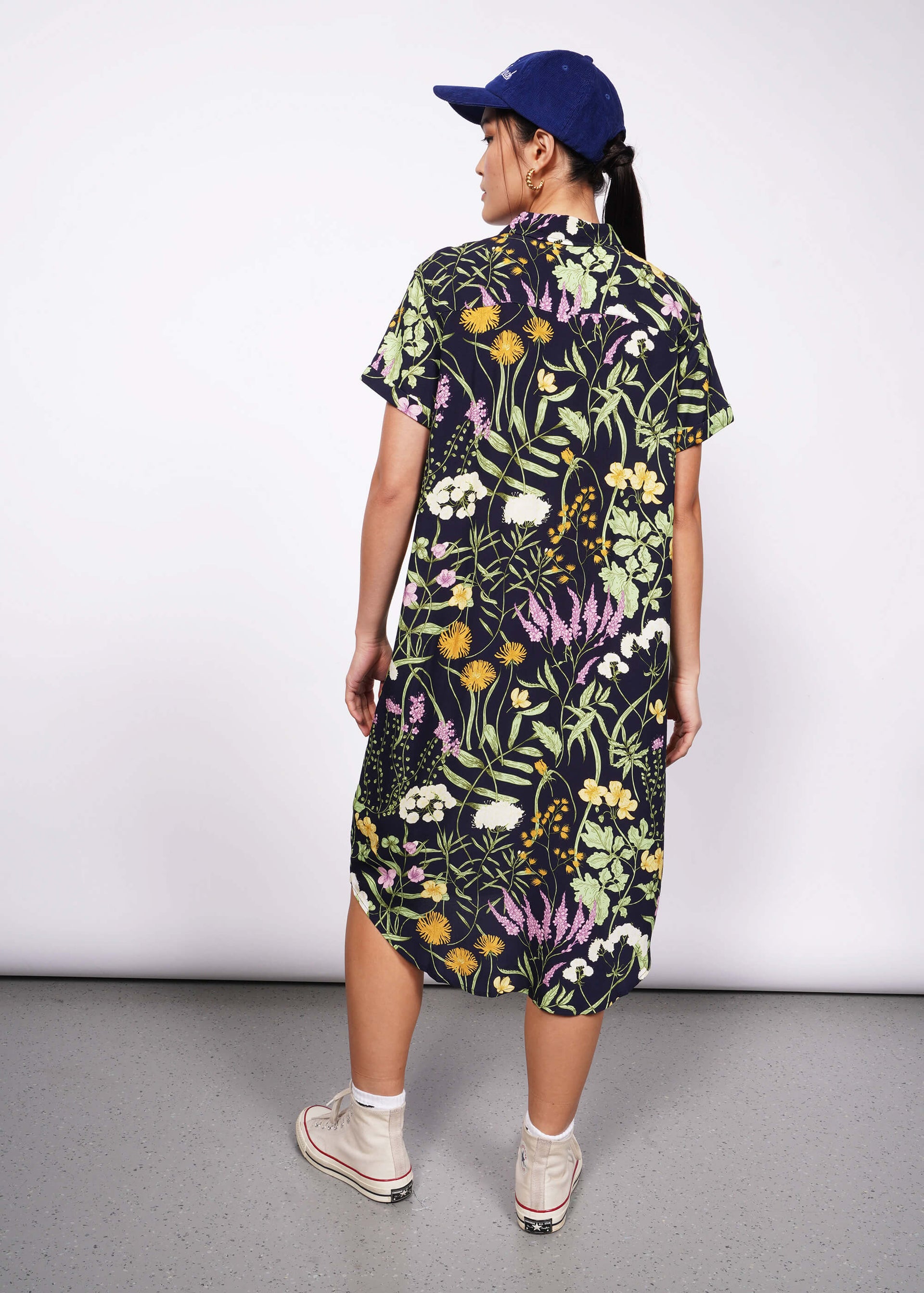 Back view of person with hand on hip looking at camera wearing botanical printed short sleeve button up shirt dress, Converse, and baseball cap.