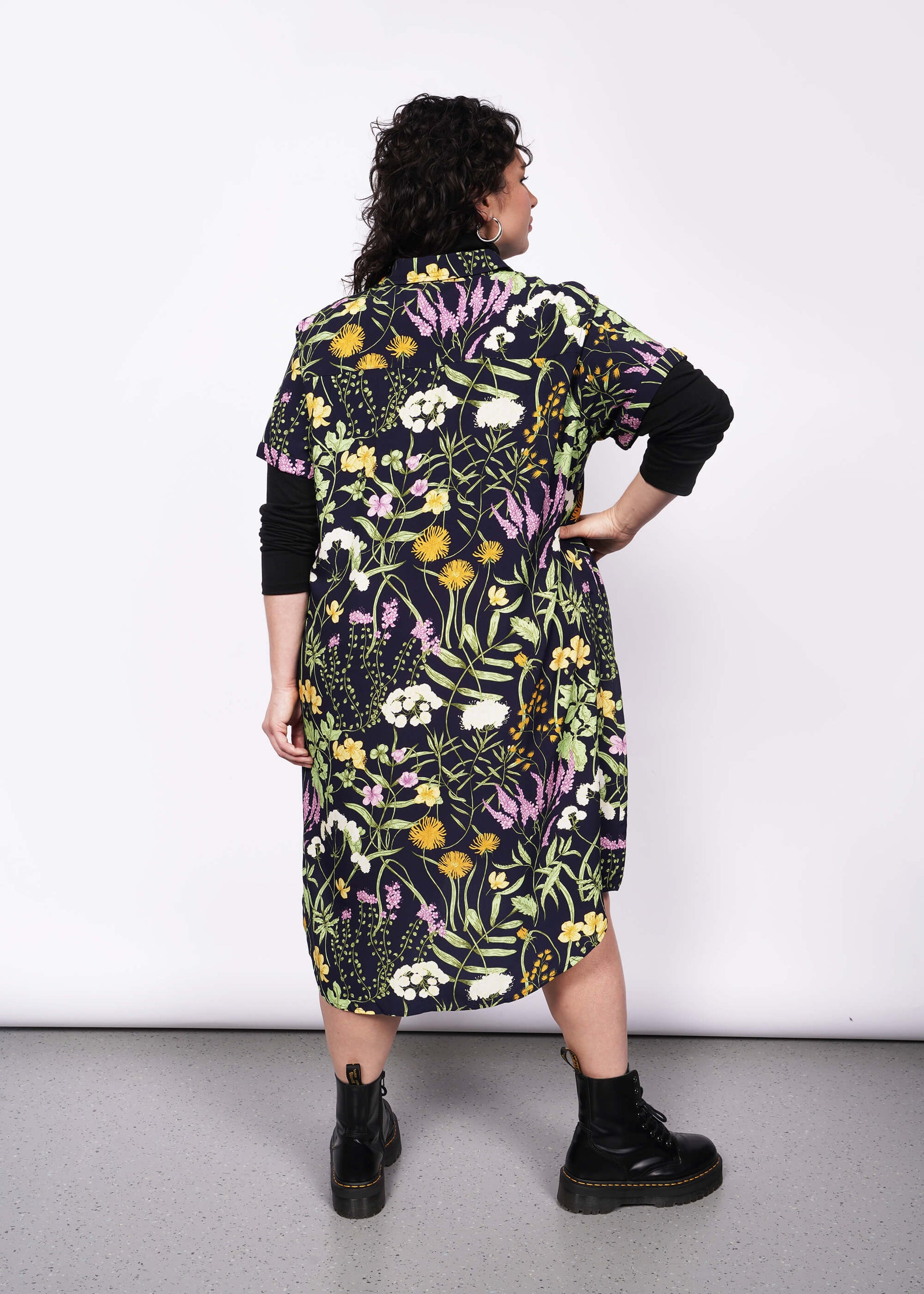 Back view of person with short curly black hair looking at camera wearing black long sleeve turtleneck layered under short sleeved botanical printed shirt dress and Doc Martens boots.