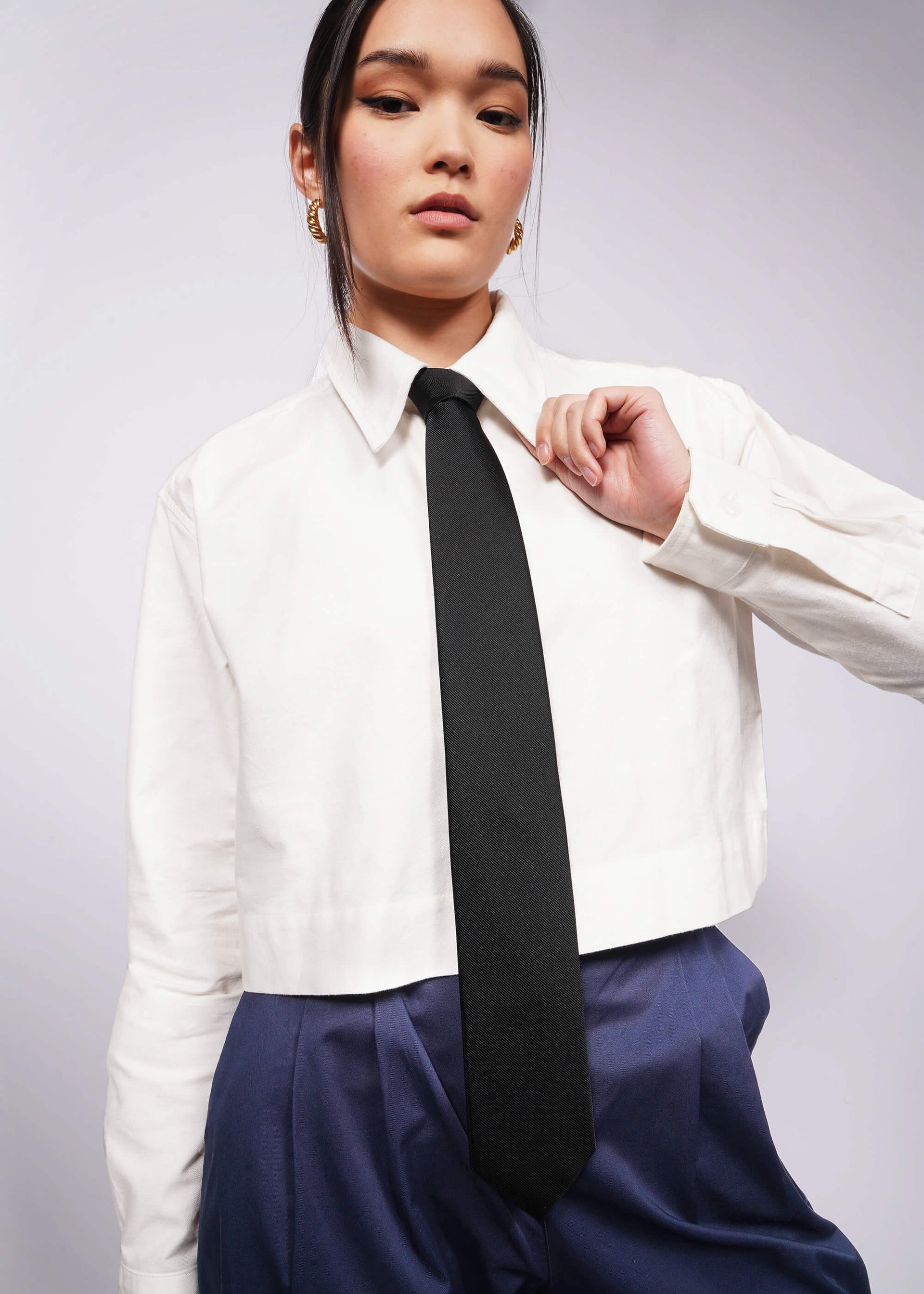 Person with black hair tied back wears cropped white button up, navy pants, and black silk tie.