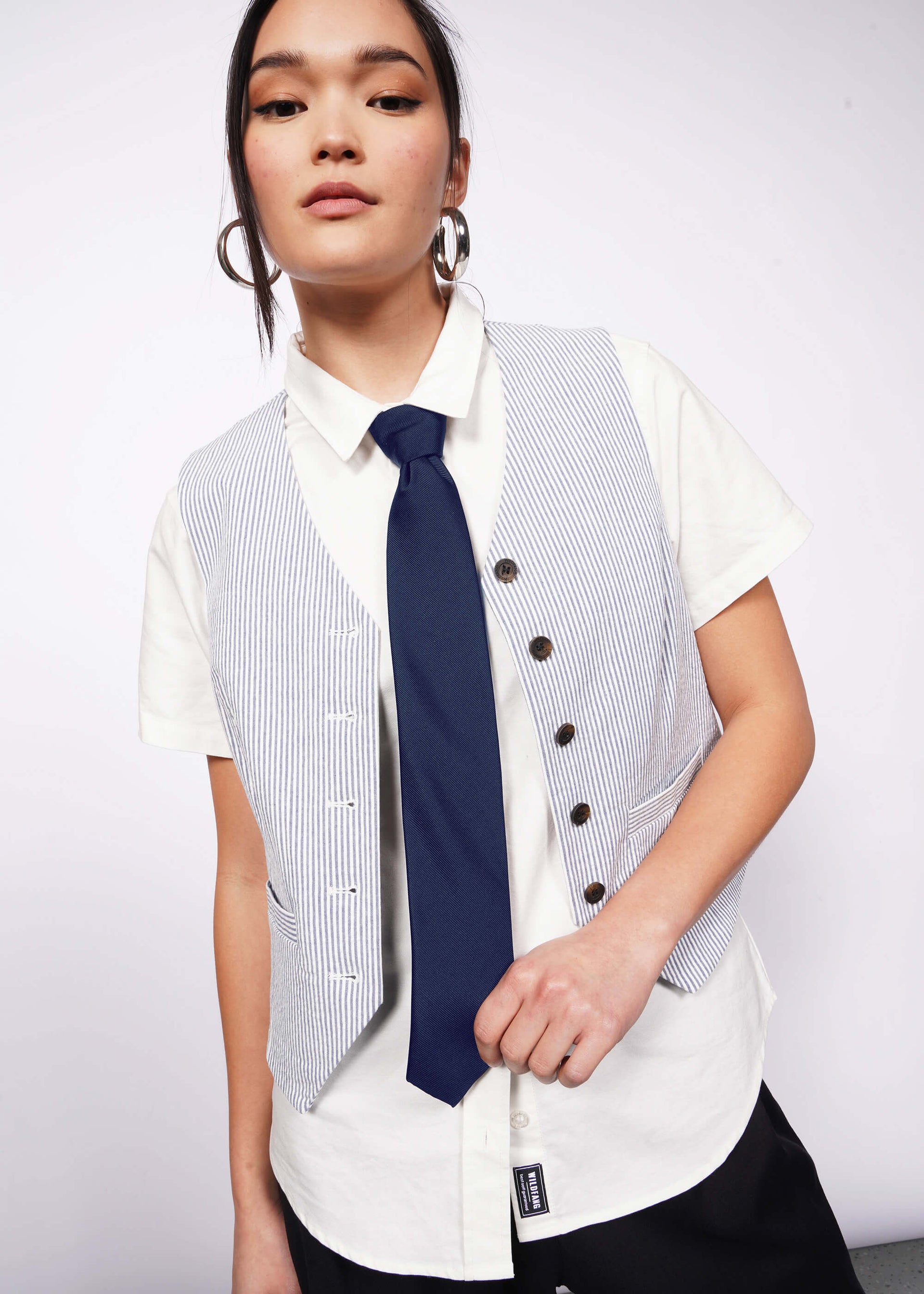 Model with black hair wears silver hoop earrings, white short sleeve button up, seersucker vest unbuttoned, and navy blue tie.