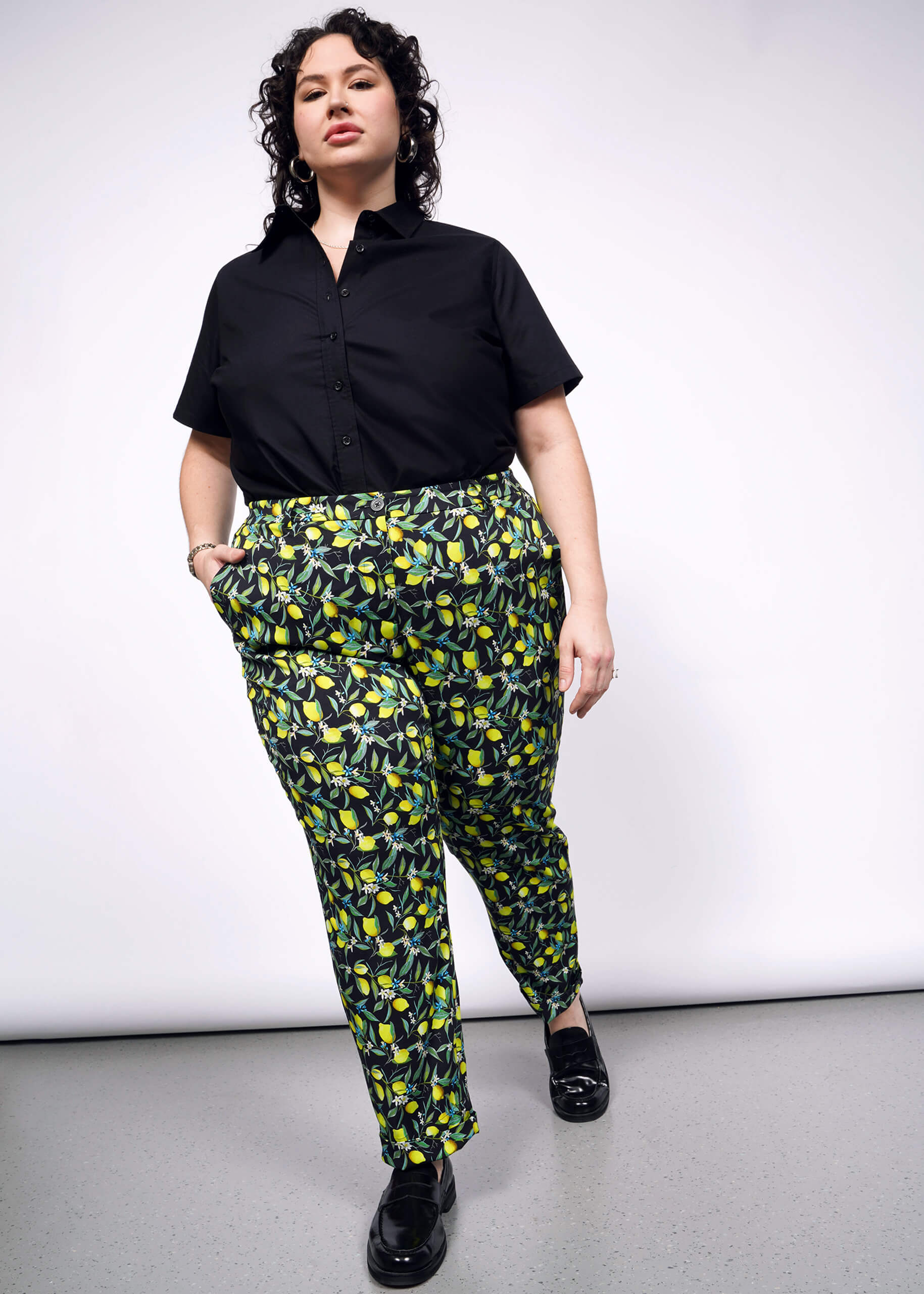 Person wearing a black shirt and lemon-patterned pants walks forward on a gray floor against a white background.