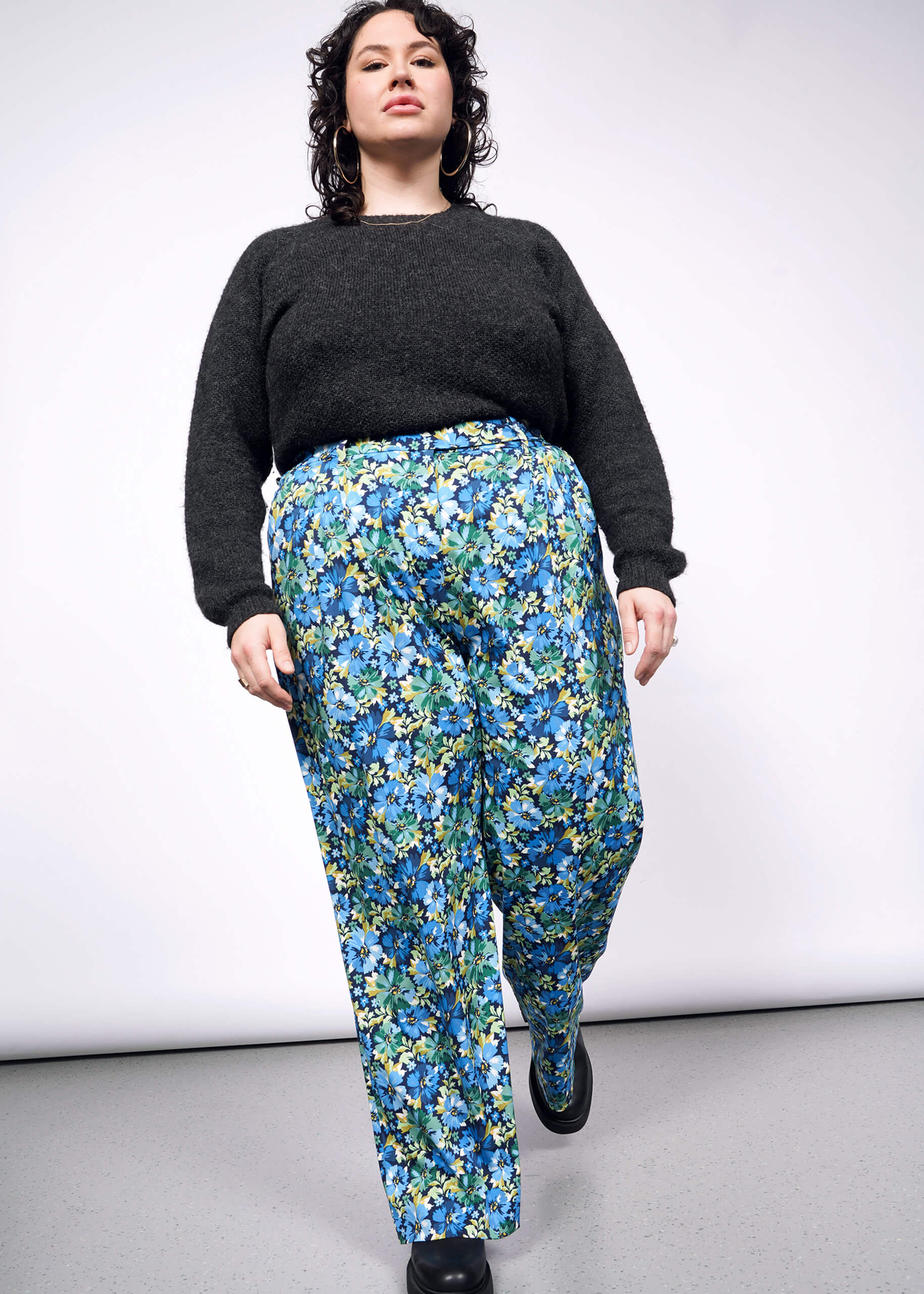 Person with curly short black hair looks at camera while wearing black sweater tucked into patterned floral green and blue wide leg pants and black boots.