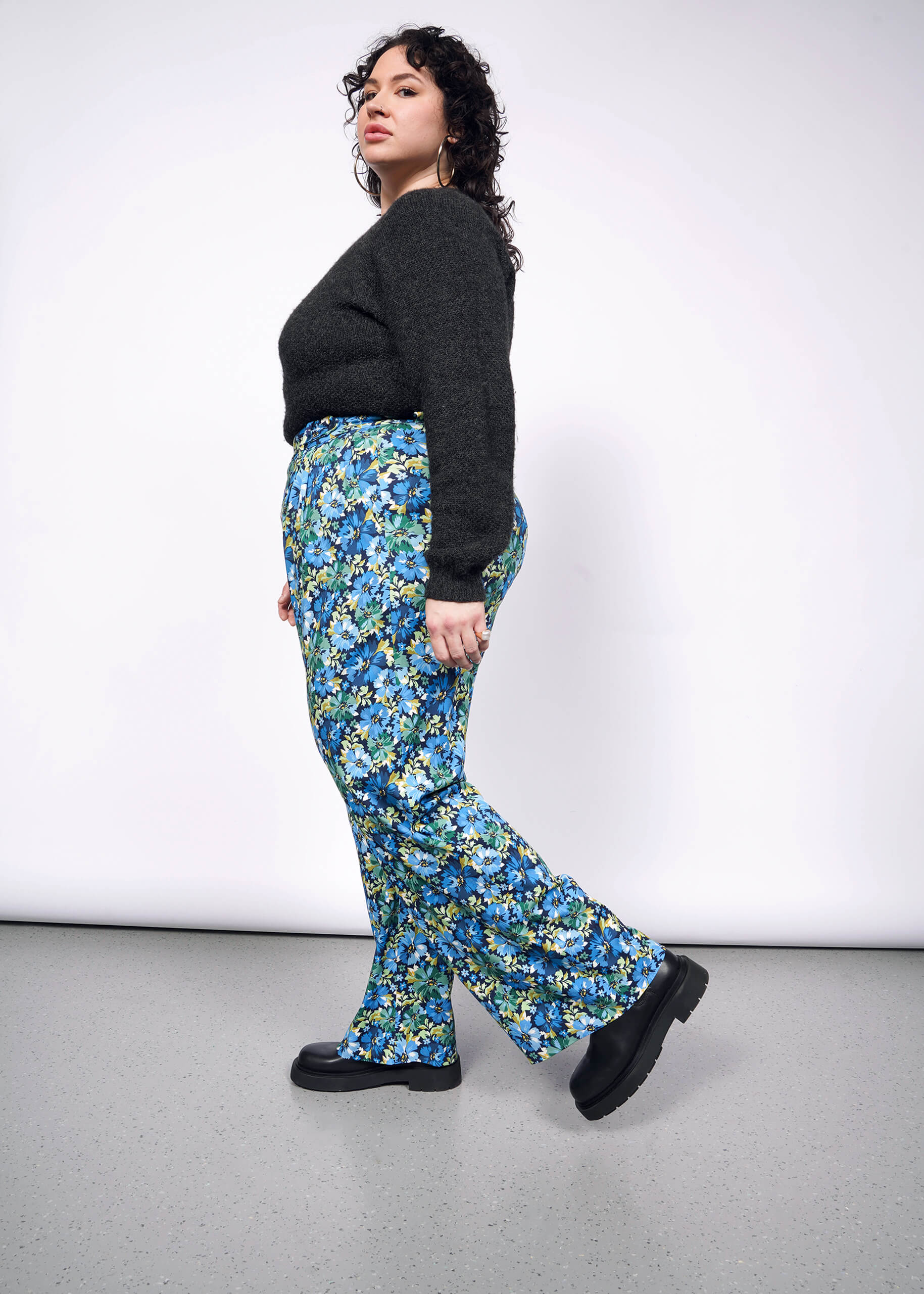 Side view of person with curly short black hair looking at camera wearing black sweater tucked into patterned floral wide leg trousers and black boots.