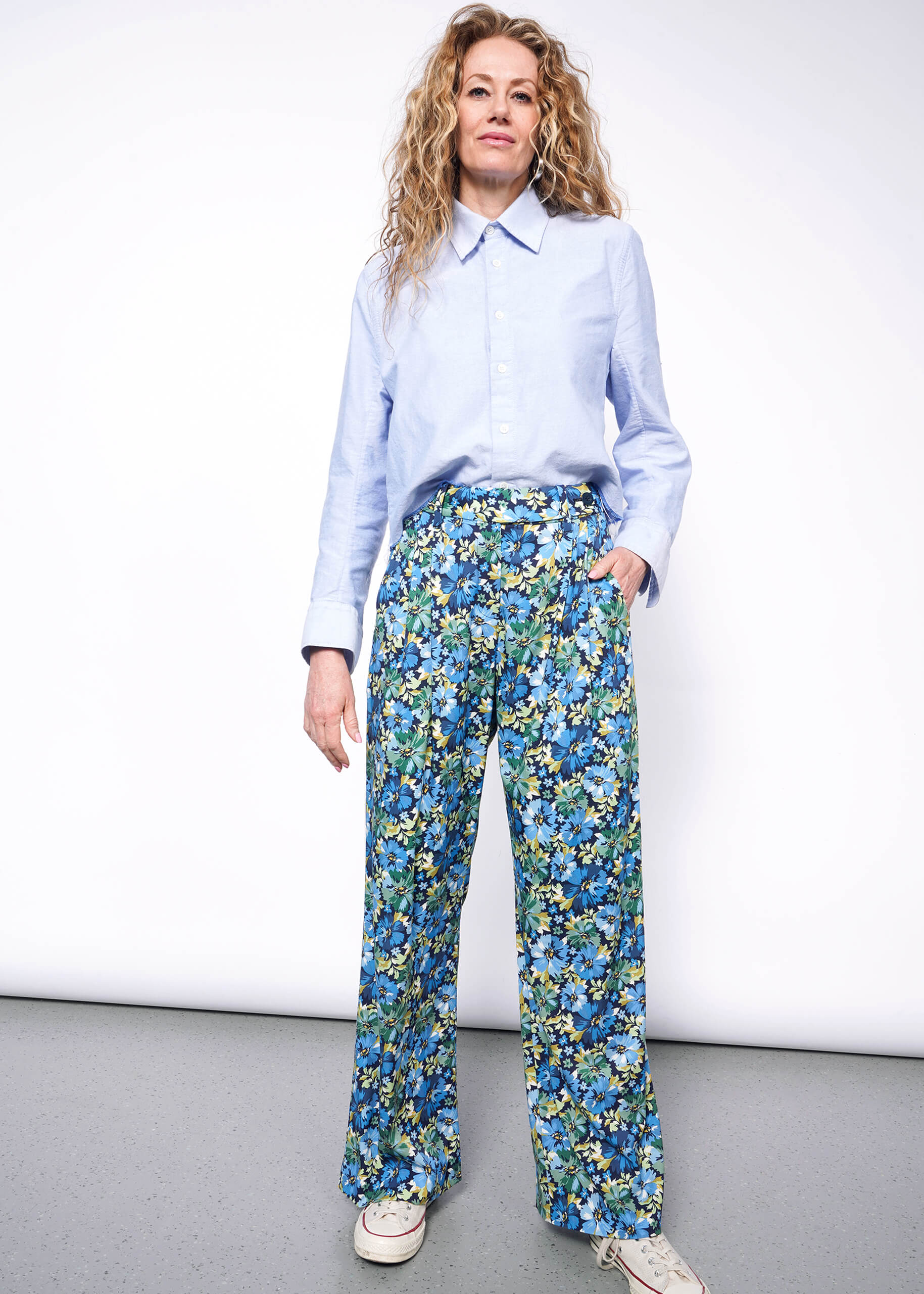 Person with medium length wavy blonde hair looks at camera wearing light blue button up shirt tucked into patterned floral blue and green wide leg trousers and Converse sneakers. Neutral background.