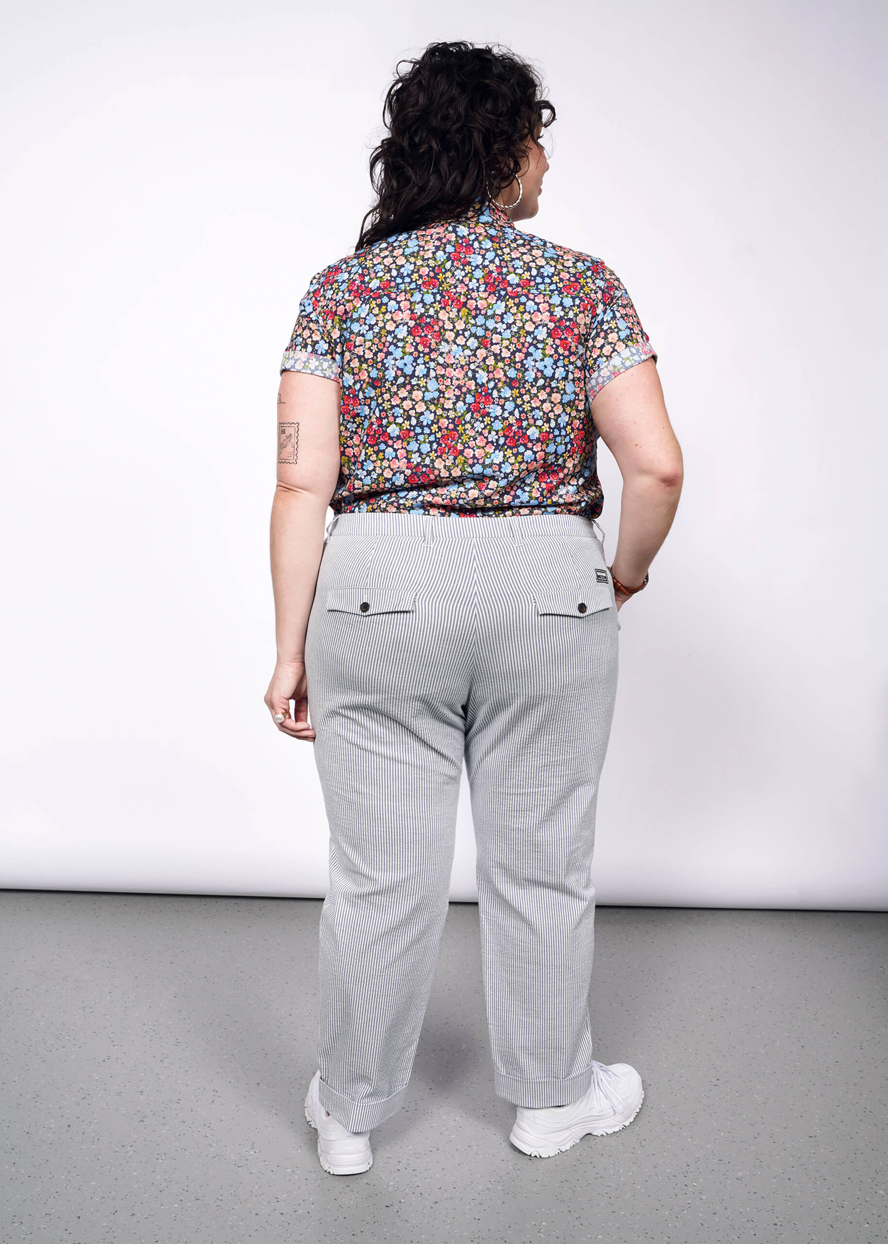 Back view of person with short black curly hair wearing seersucker pants, white sneakers, and flower printed short sleeve button up shirt.