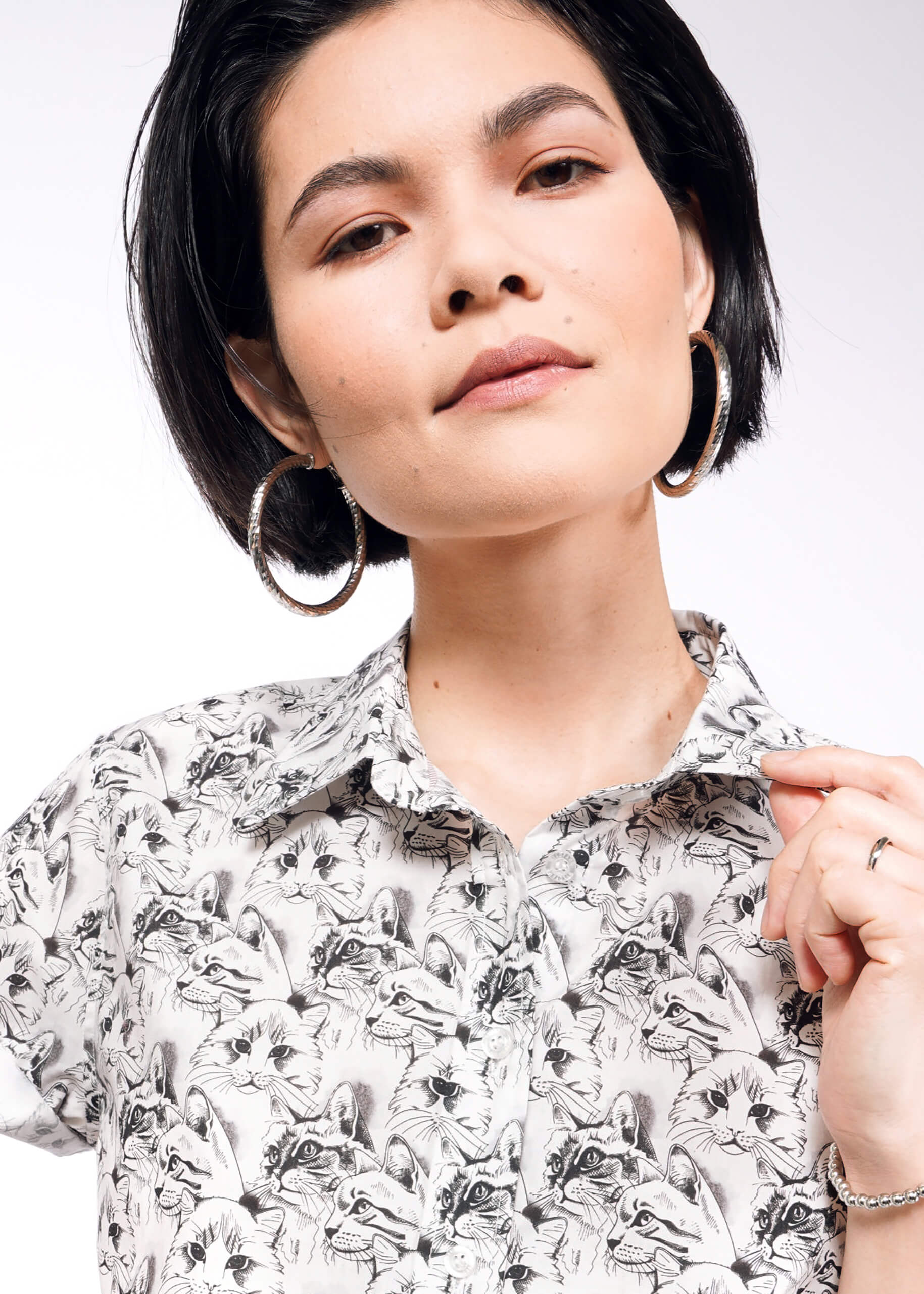 A person with short dark hair wears large hoop earrings and The Essential Button Up, a bold patterned shirt featuring animal illustrations. It has a tailored fit with Wildfang branded buttons for unique flair. They look calmly at the camera against a white background.