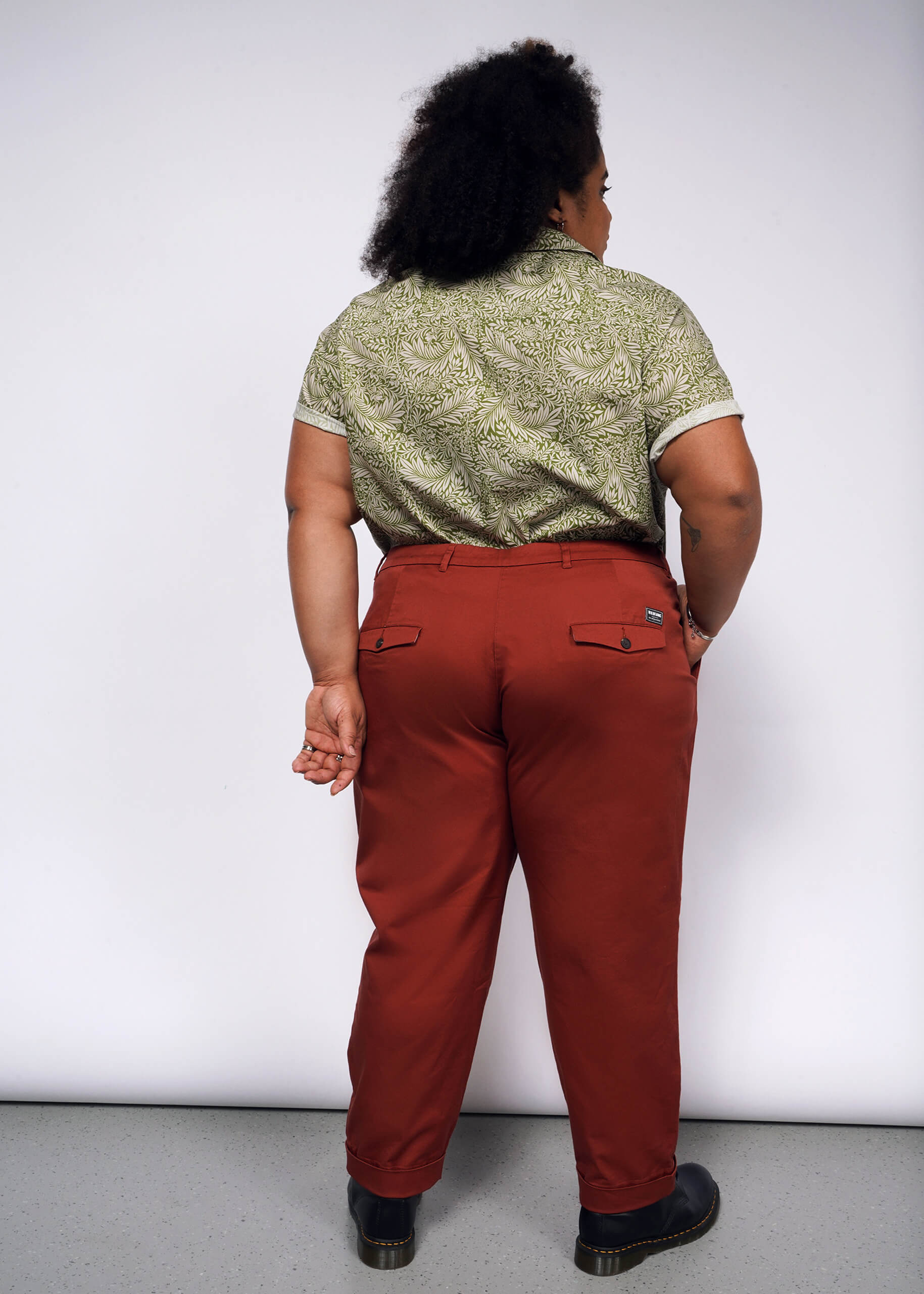 Against a white background, a person with curly hair wears The Essential Button Up, featuring a vibrant Wildfang classic green pattern in breathable cotton, paired with rust-colored tailored trousers and black shoes. One hand is elegantly positioned behind their back.