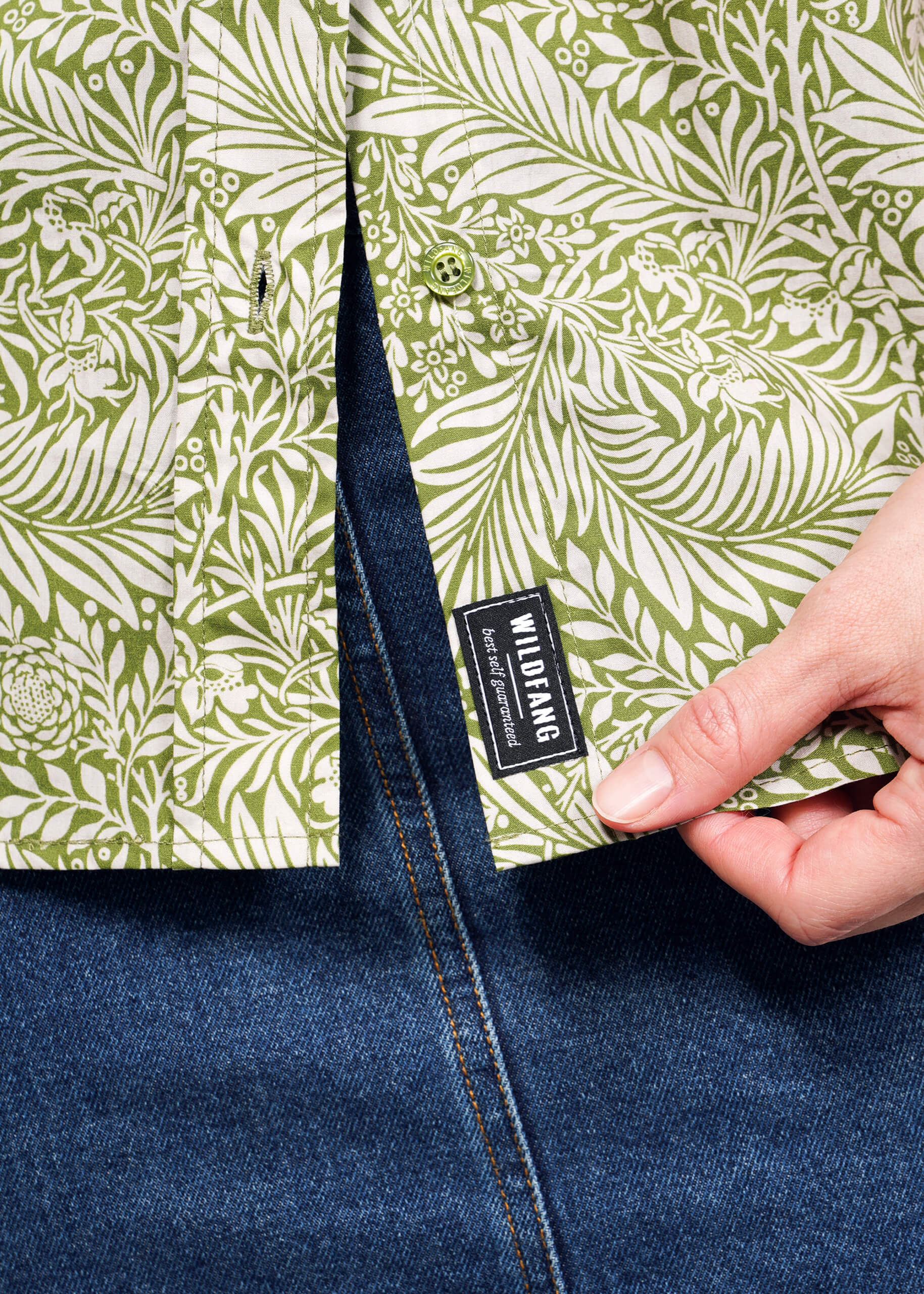 A person holds the corner of The Essential Button Up by Wildfang, showcasing its green and white botanical pattern. Made from breathable cotton, it is paired effortlessly with blue denim jeans.