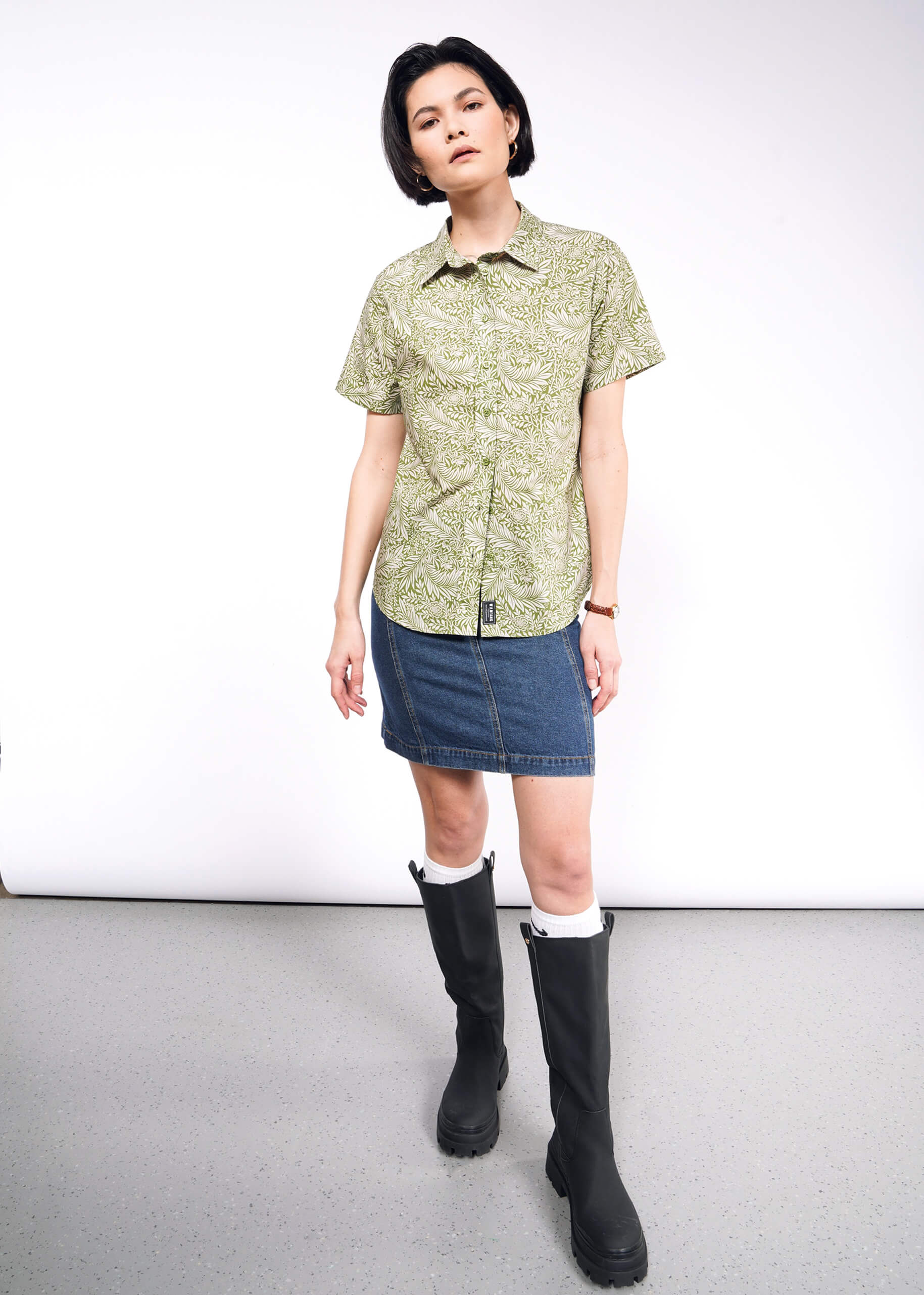 A person with short black hair wears The Essential Button Up in a green patterned tailored fit, paired with a denim skirt and black knee-high boots. They stand against a white background, looking forward with relaxed ease.