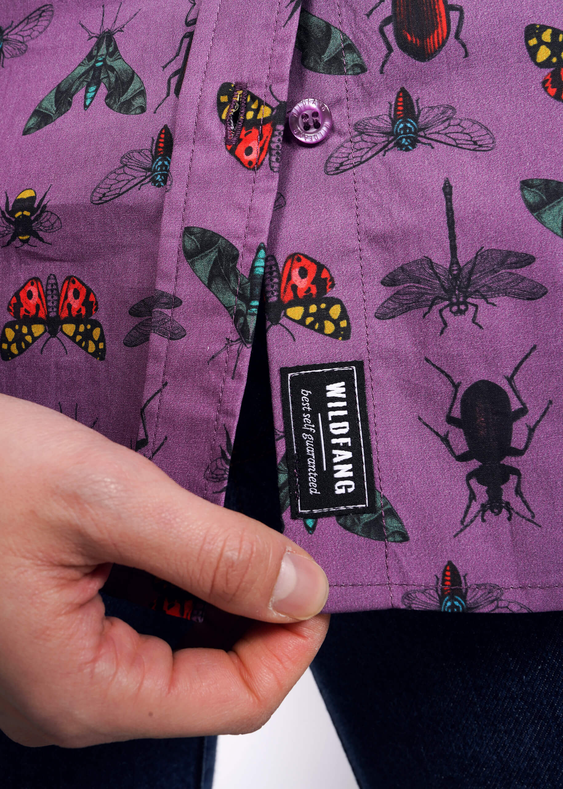 Close up view of model holding bottom hem of purple bug printed button up showing WILDFANG best self guaranteed label. 