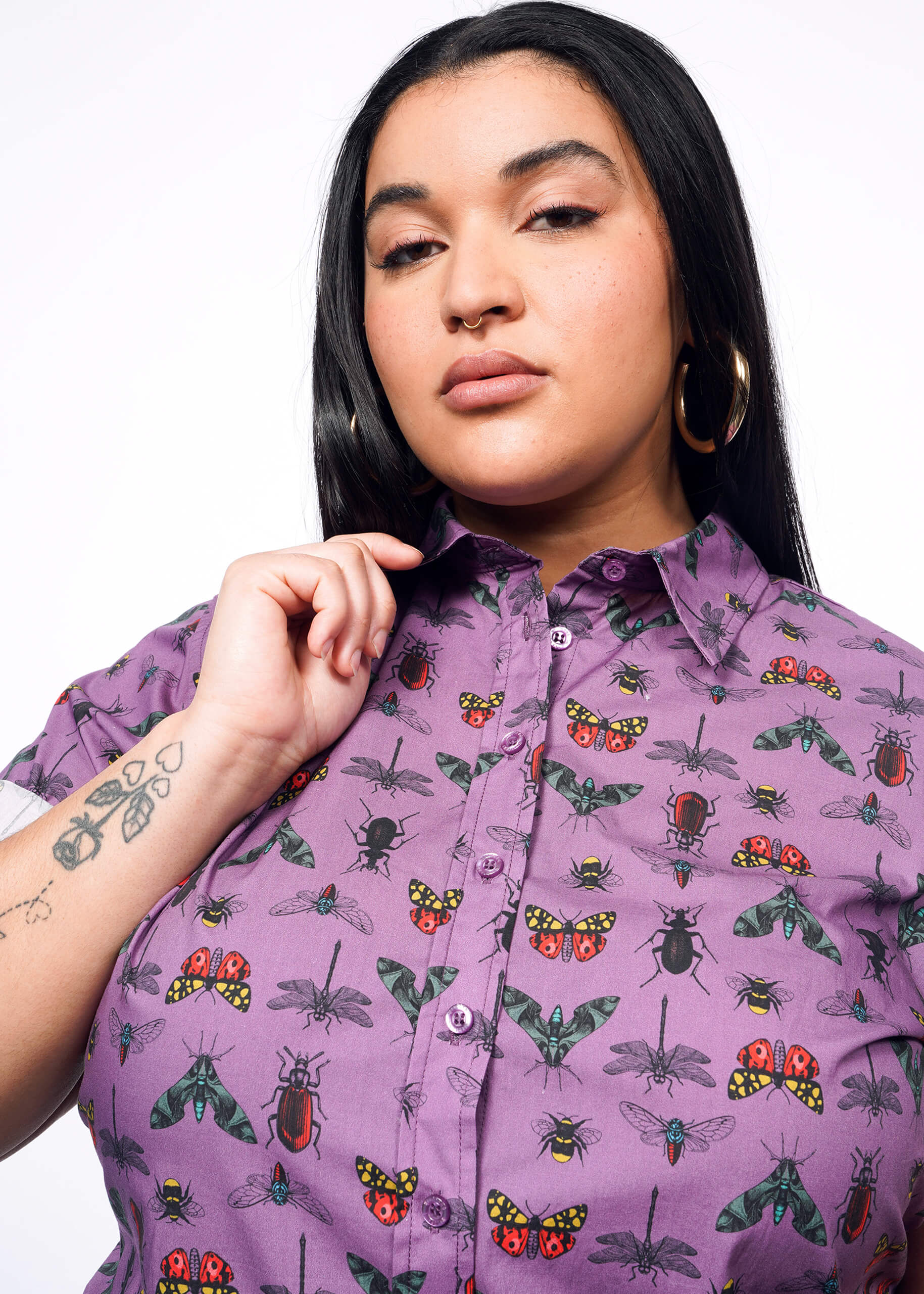 Close up of person with long black hair, nose ring wearing hoop earrings and holding lapel of purple bug printed button up.