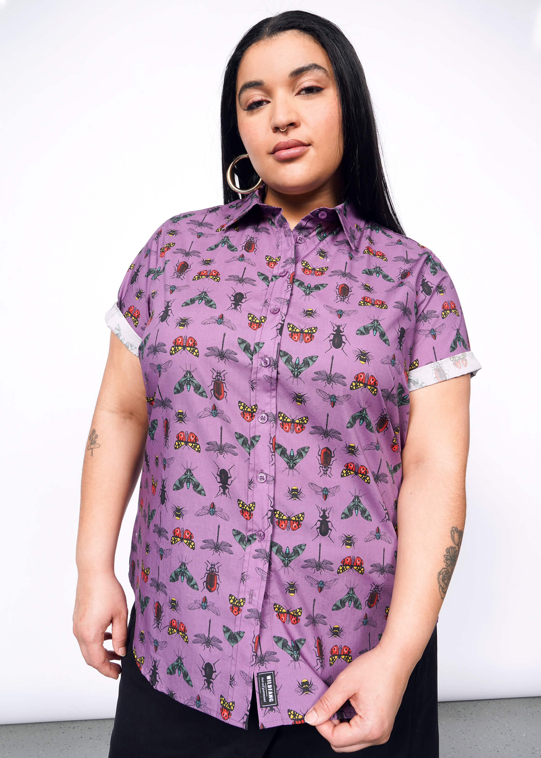 Person with long black hair and nose ring is wearing hoop earrings and holding bottom of purple bug printed button up.